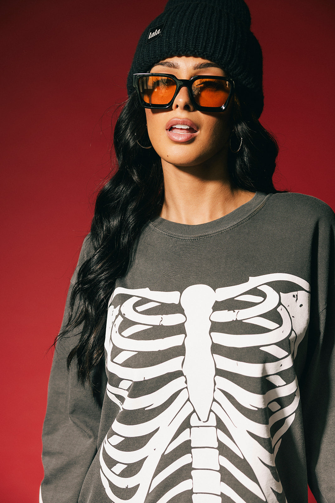 Person wearing a dark long-sleeve shirt with a skeleton design, black beanie, and orange sunglasses against a red background.