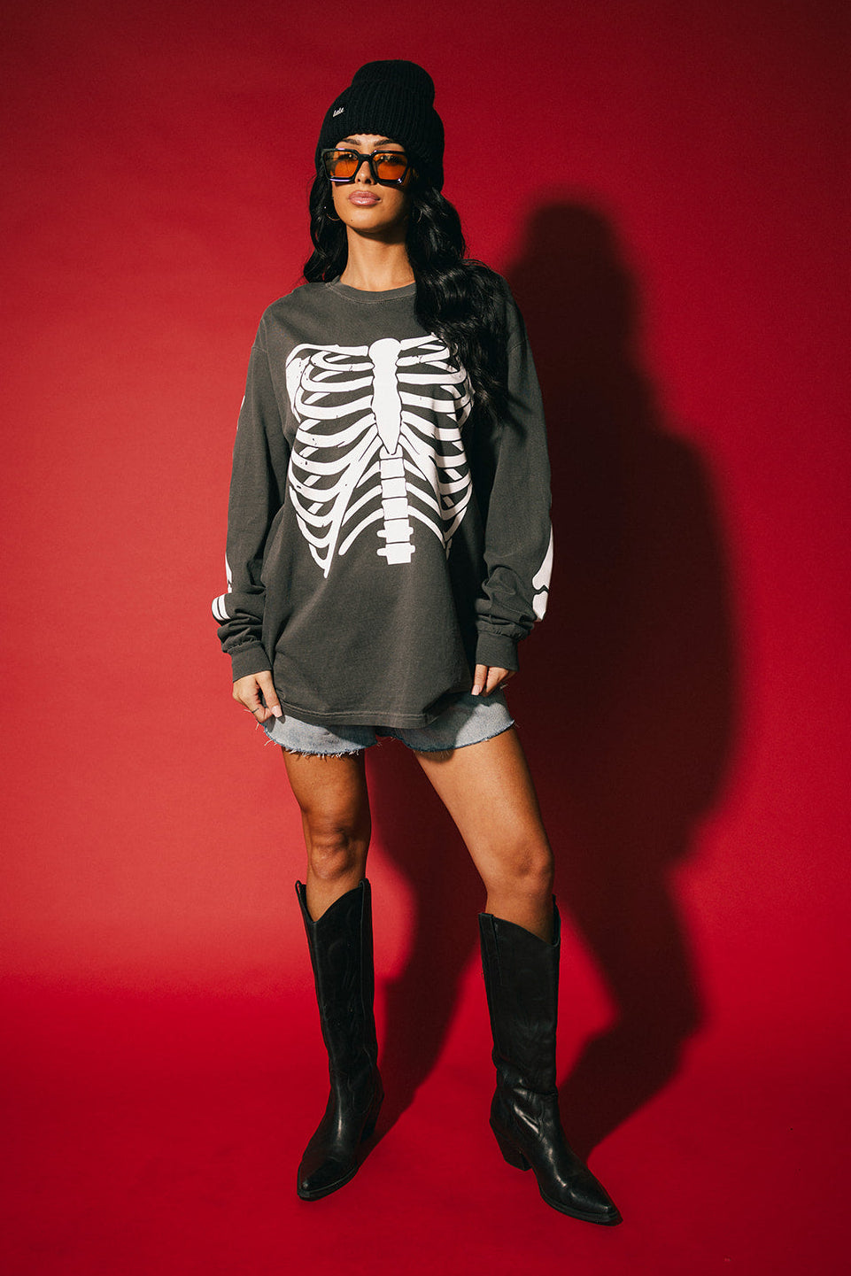 Person wearing a dark gray sweatshirt with a skeleton design, denim shorts, black knee-high boots, and a black beanie on a red background.