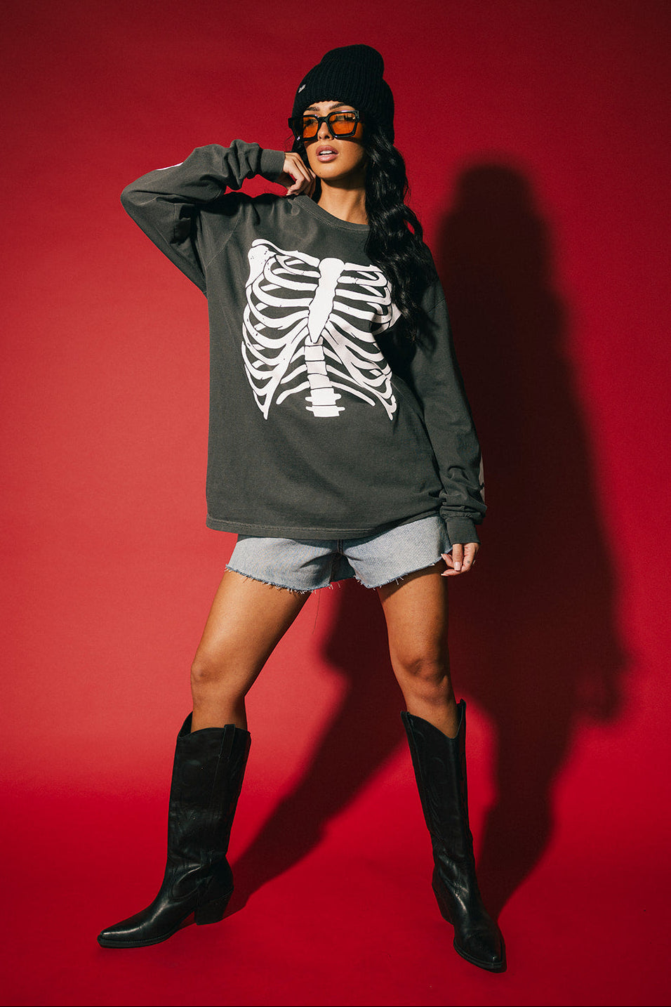 Person wearing a dark gray sweatshirt with a skeleton design, shorts, and black boots on a red background