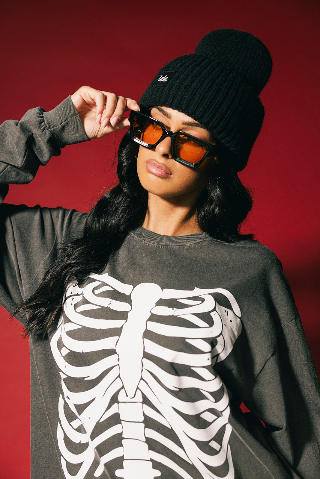 Person wearing a dark gray sweatshirt with a skeleton design, black beanie, and orange sunglasses against a red background.