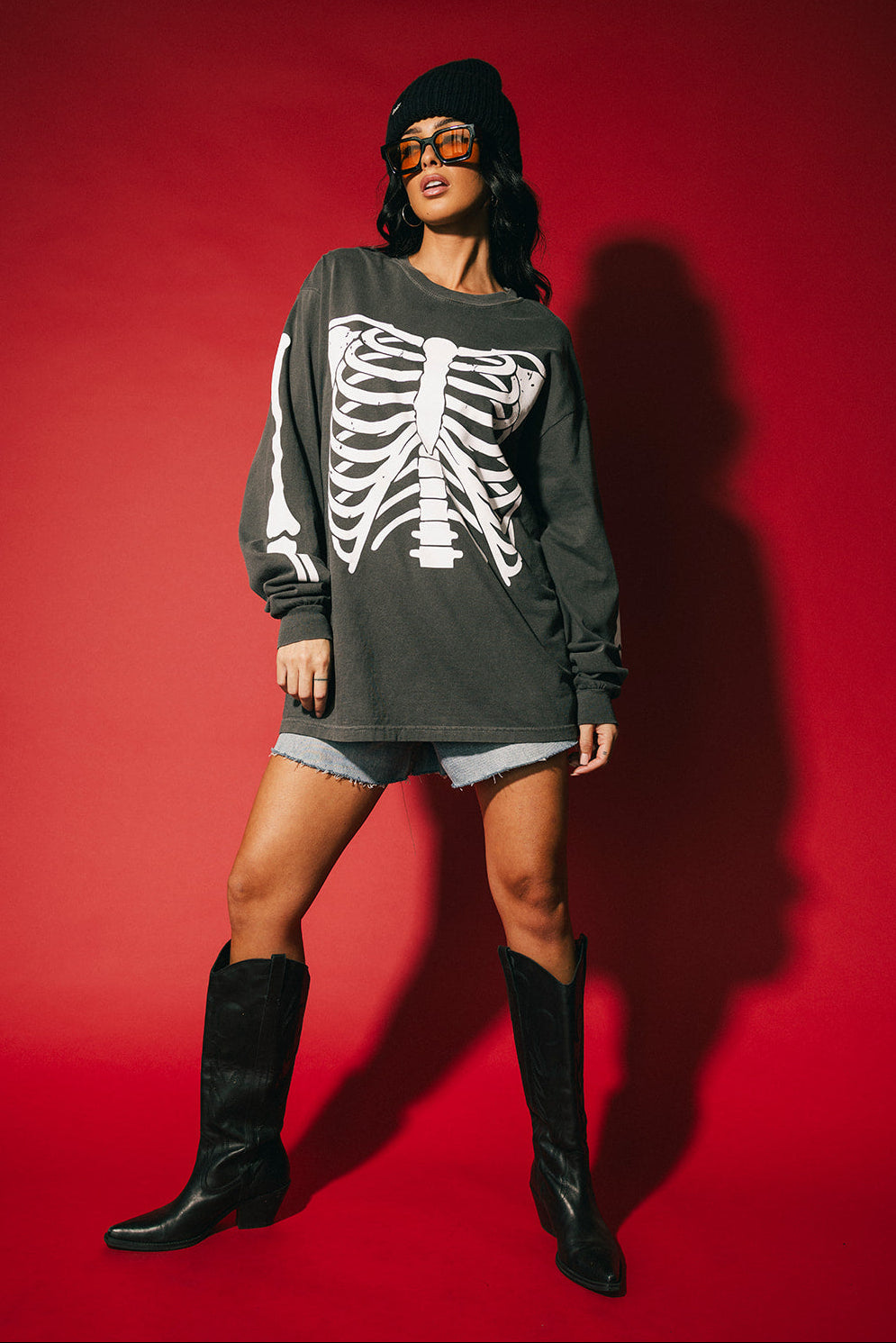 Person wearing a dark long-sleeve shirt with a skeleton design, shorts, and black boots against a red background.