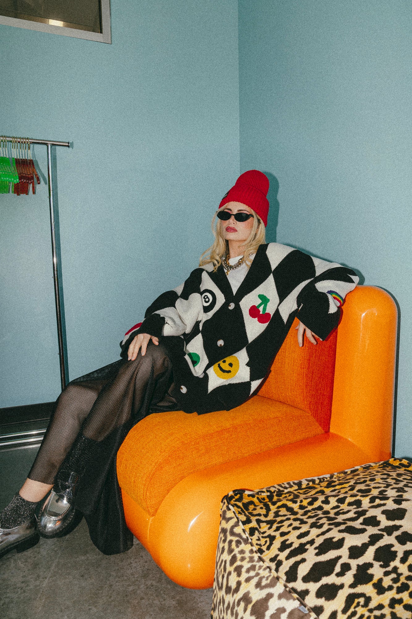 Person sitting on an orange chair wearing a black and white patterned coat with a red beanie in a room with light blue walls.