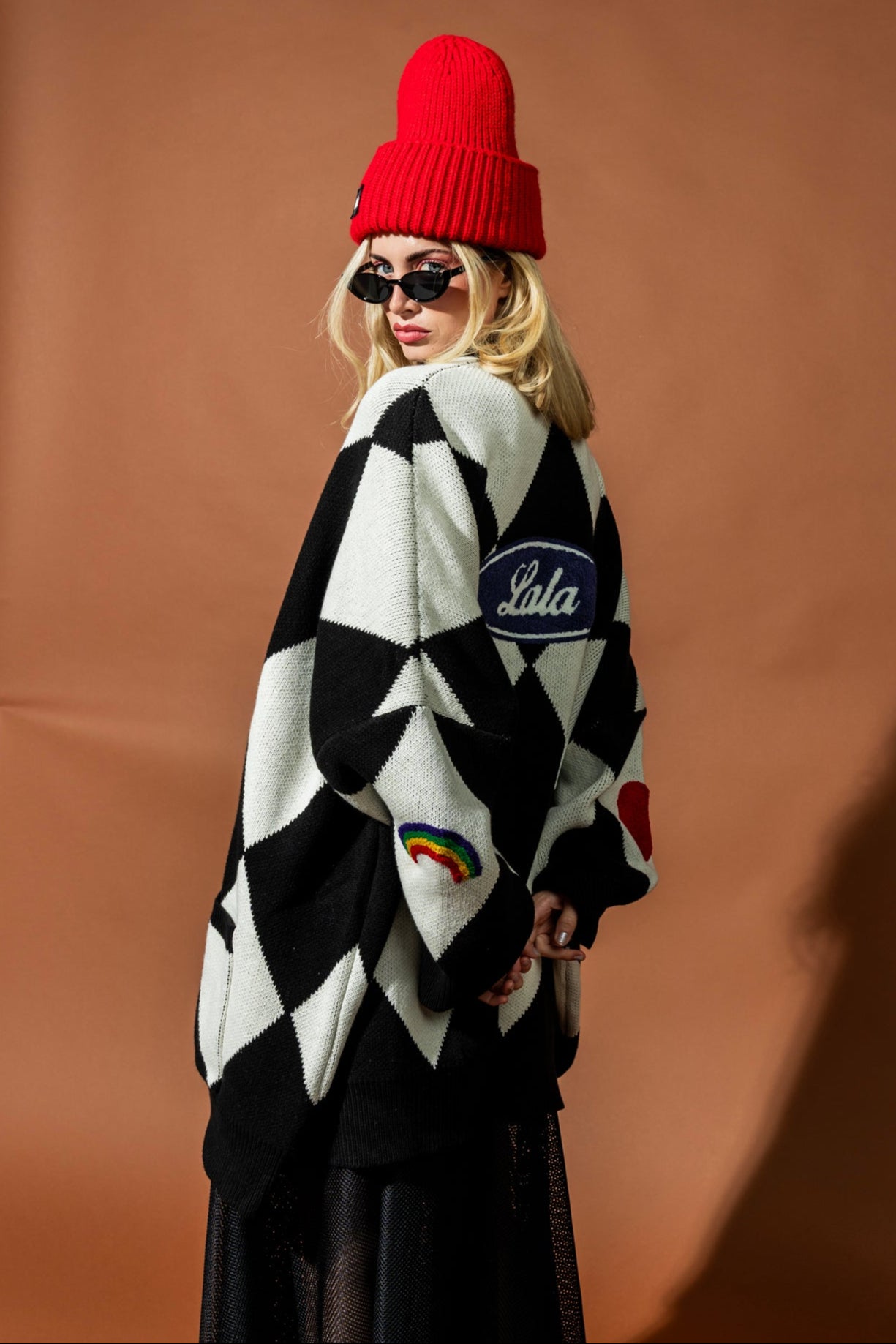 Person wearing a black and white patterned cardigan with 'Lala' branding, red beanie, and sunglasses on a brown background