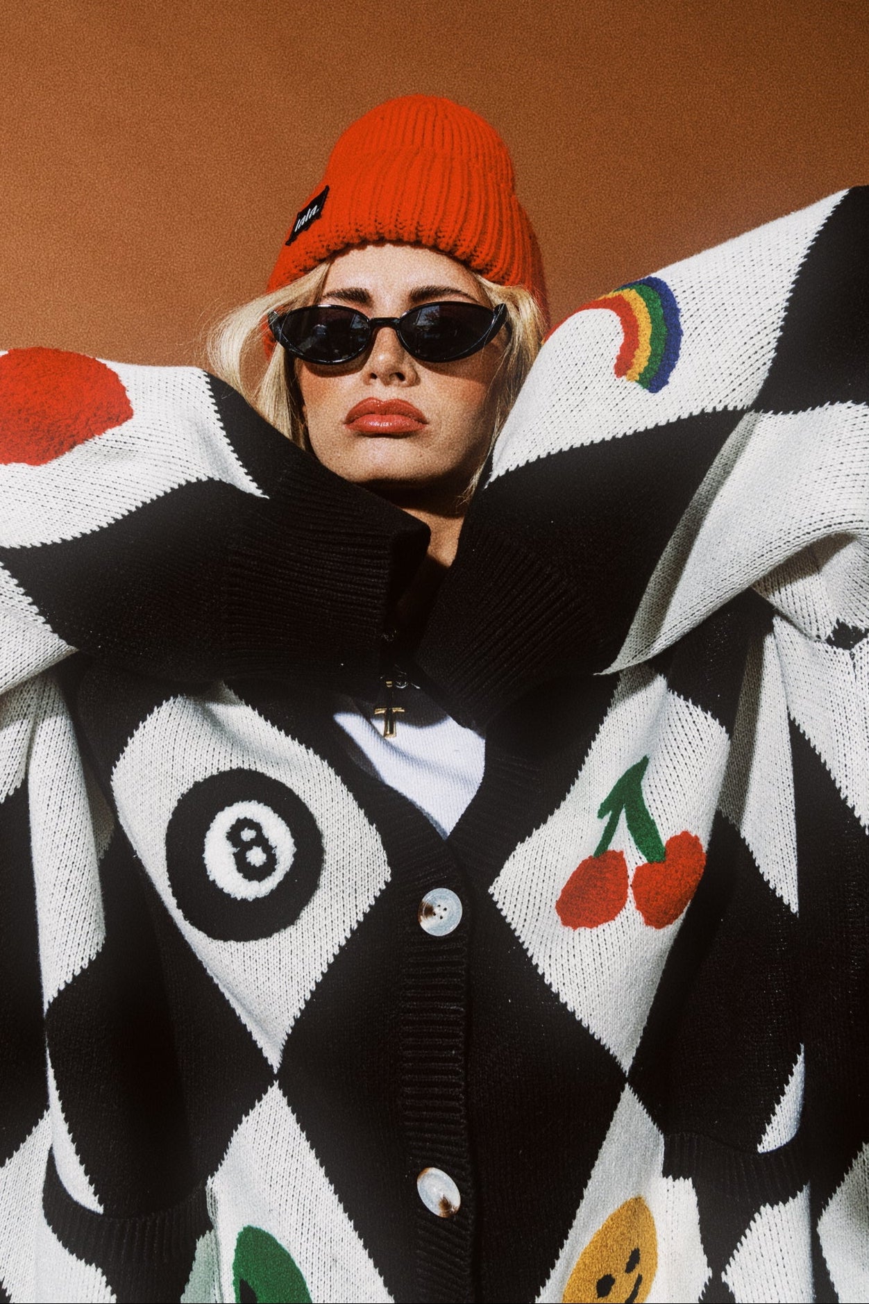 Person wearing a black and white patterned cardigan with colorful designs, sunglasses, and an orange beanie.