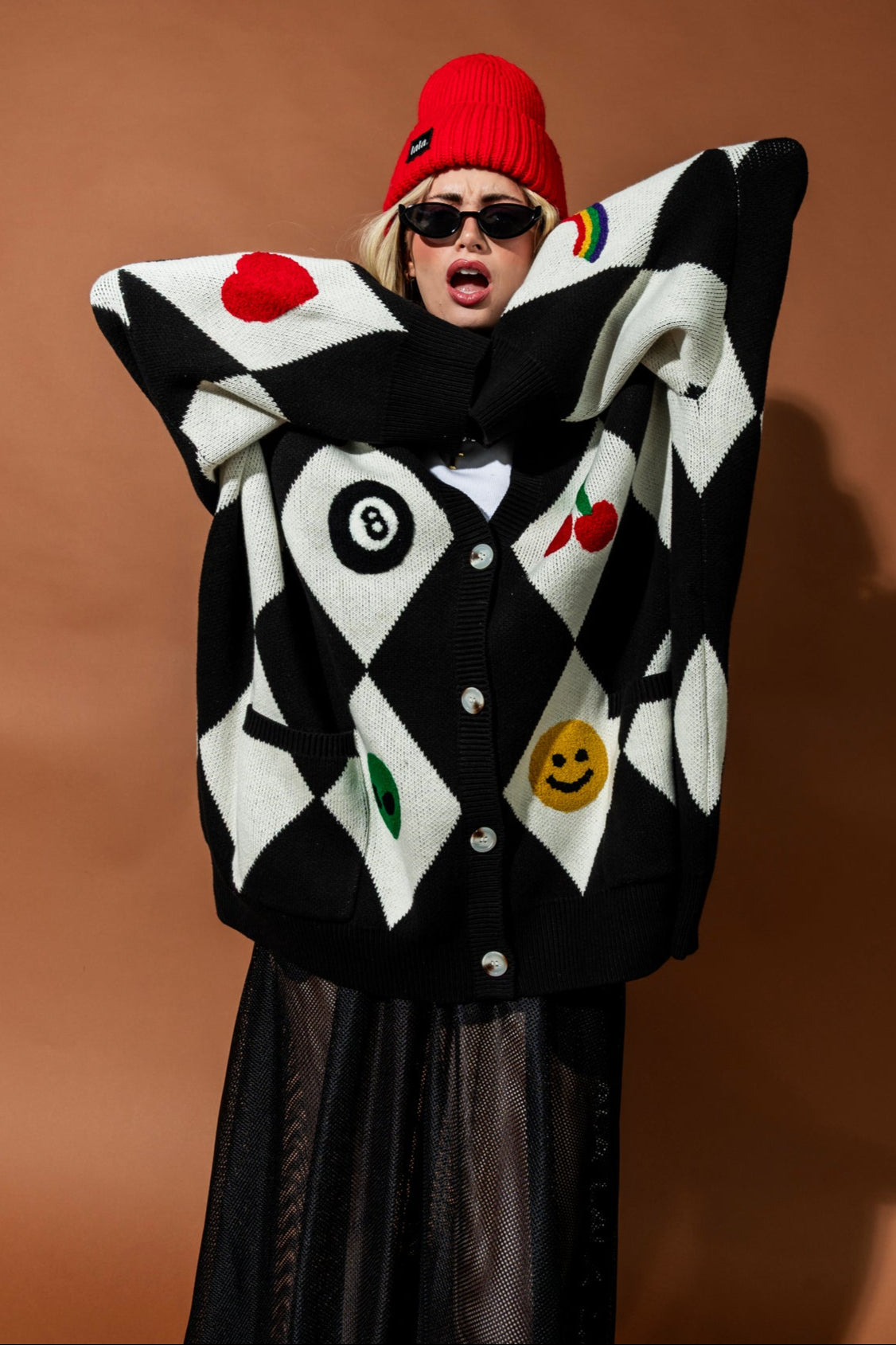 Person wearing a black and white patterned cardigan with a red beanie against a brown background