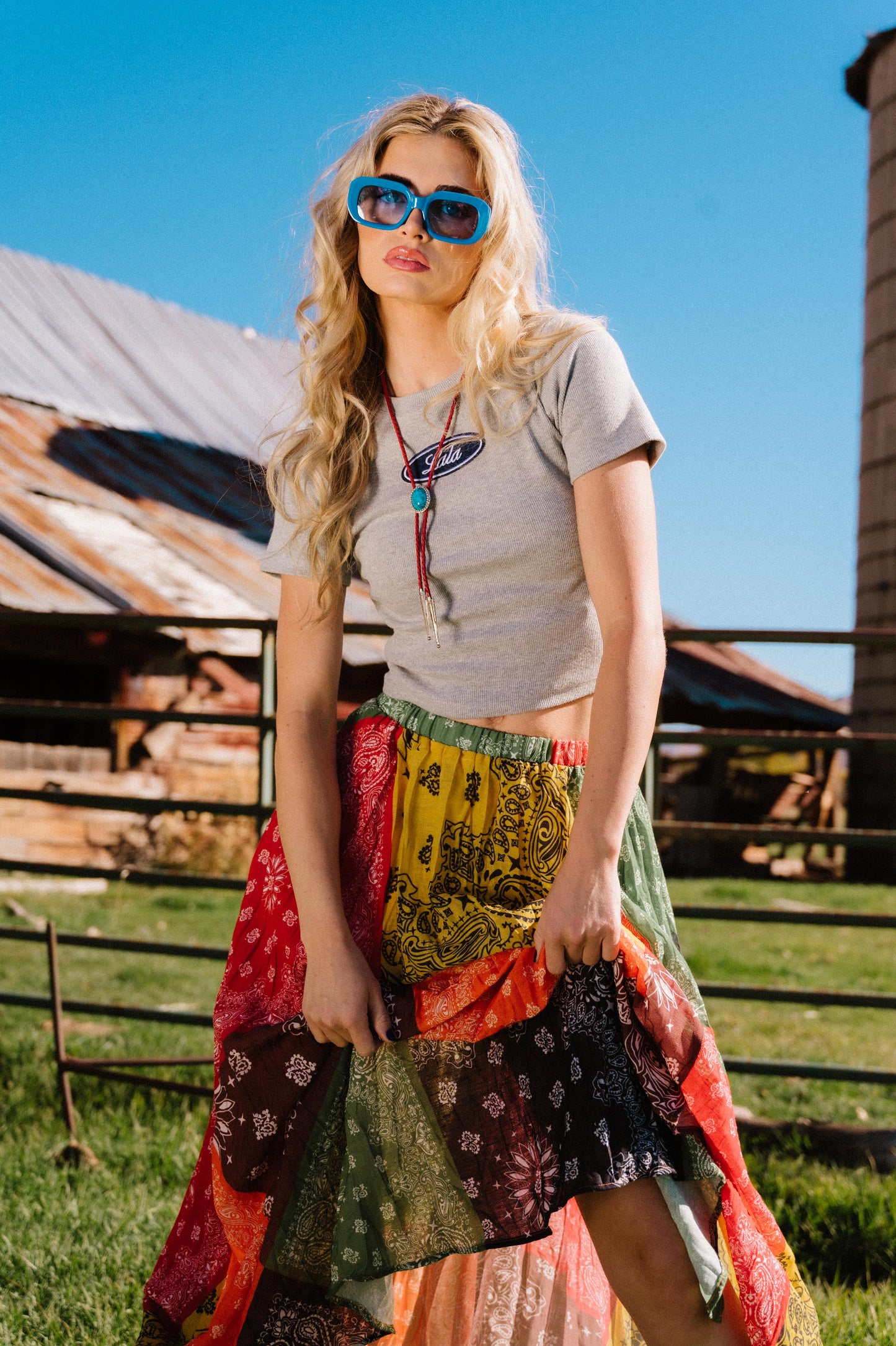 Woman wearing a grey ribbed baby tee with a 'Lala' logo, sunglasses, and a colorful skirt standing in front of a wooden barn.