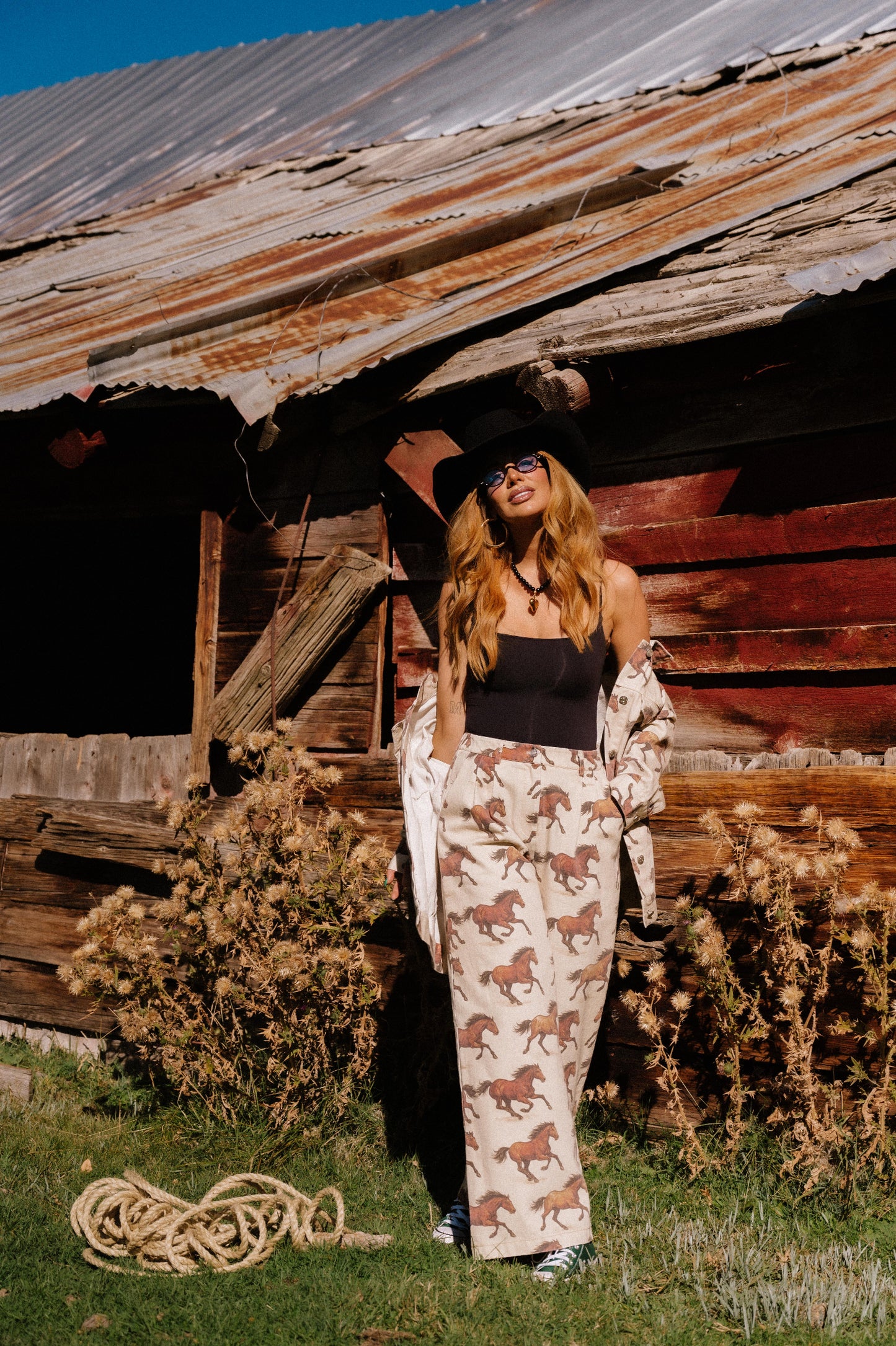 Person wearing white denim wide leg trousers with a horse pattern standing in front of a rustic wooden building with a corrugated metal roof.