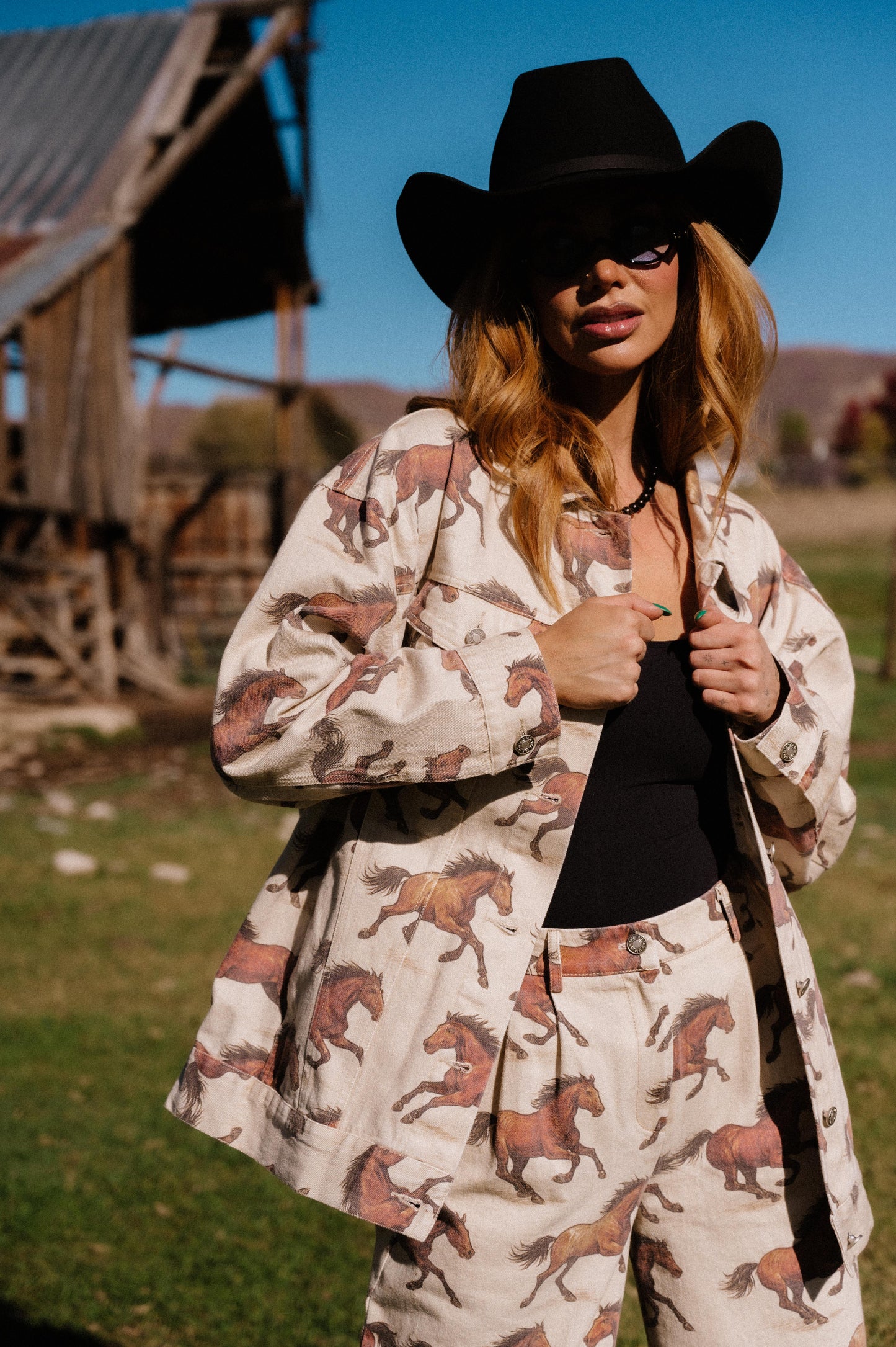 Woman wearing a cowboy hat and white denim jacket with a horse pattern in an outdoor setting