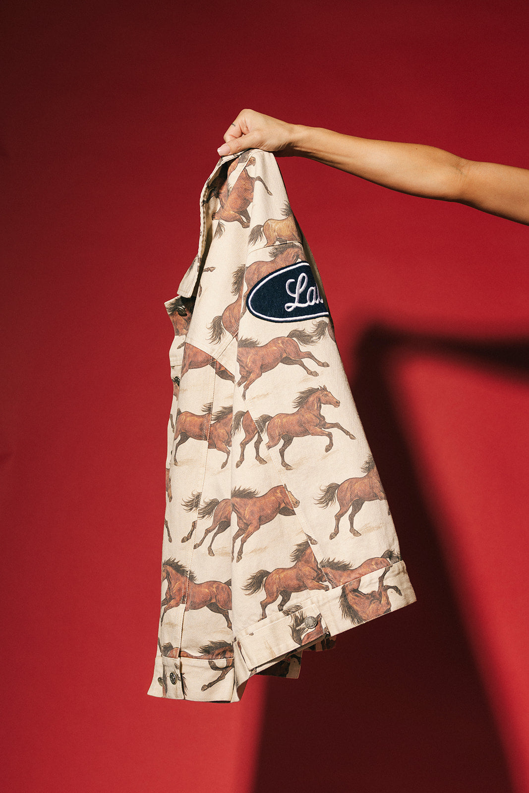 White denim jacket with a horse pattern and navy Lala label held against a red background
