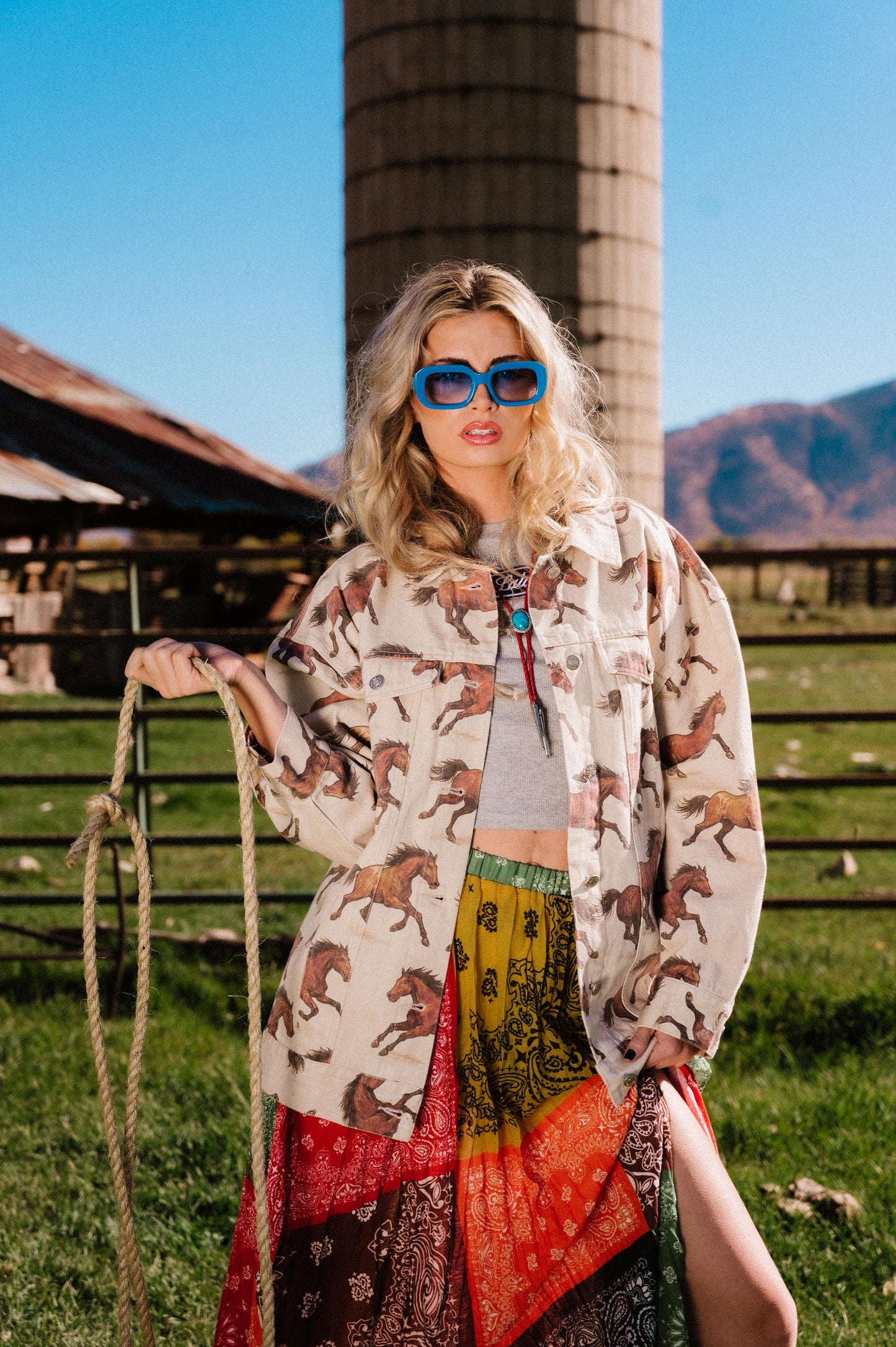 Woman in a white denim jacket with a horse pattern standing in a rural setting with mountains in the background.