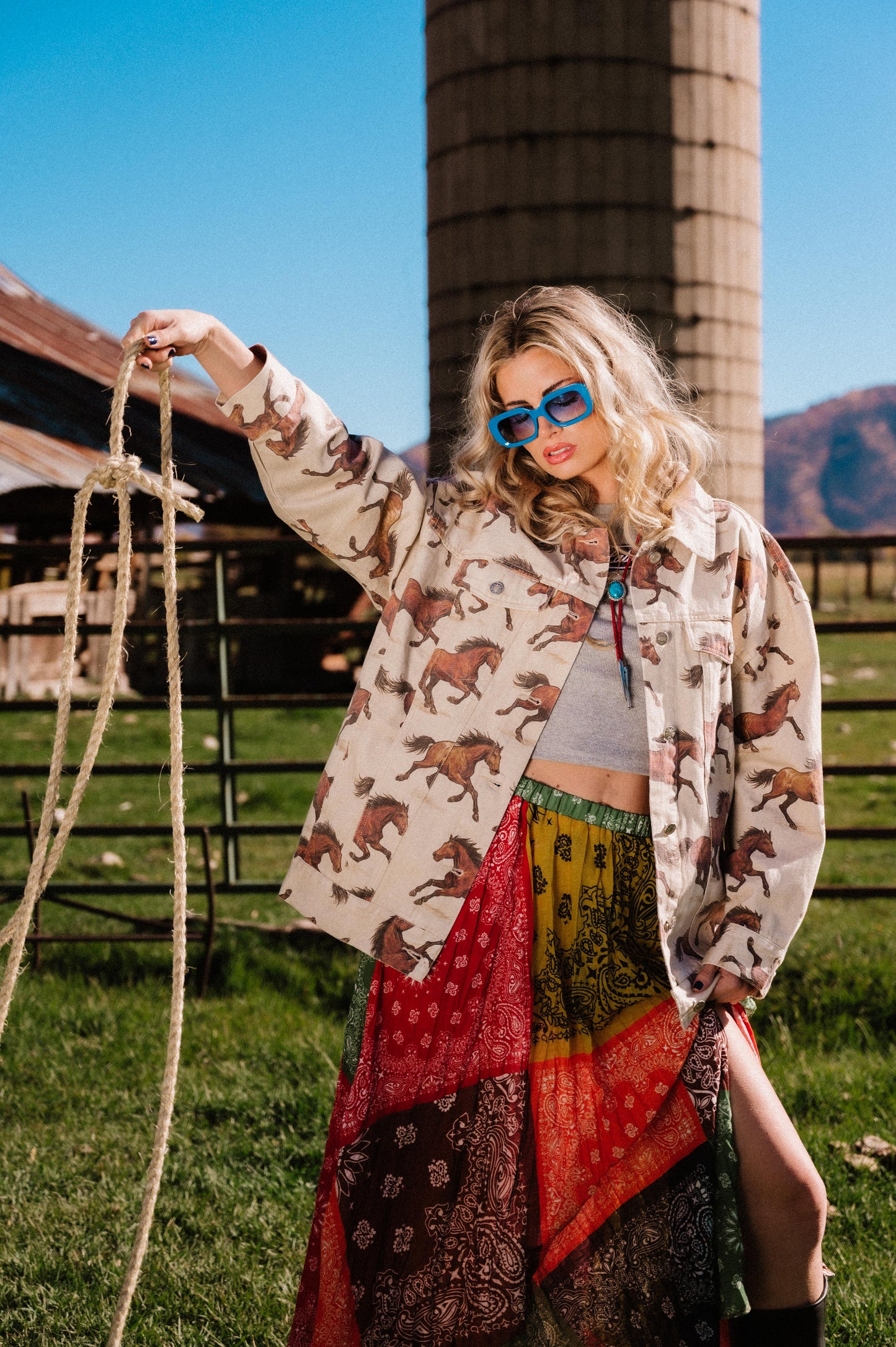 Woman in a white denim jacket with a horse pattern and colorful skirt holding a rope in a rural setting with a silo in the background.