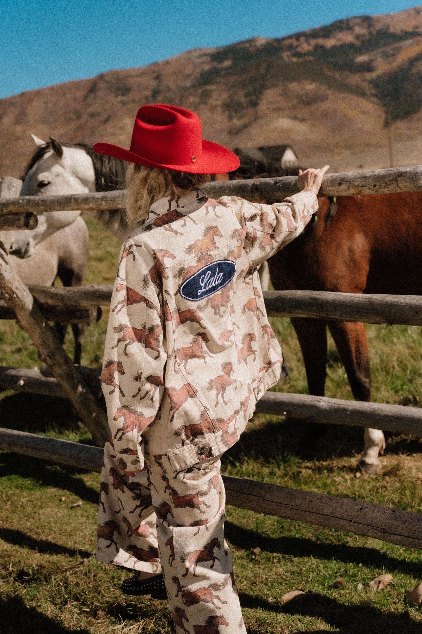 Person in white denim jacket with a horse pattern and red cowboy hat standing next to horses in a field
