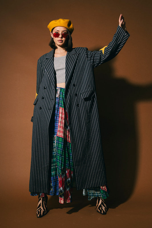Person wearing a long dark double breasted pinstripe coat with yellow star patches on the elbows, colorful skirt, and yellow beret against a brown background