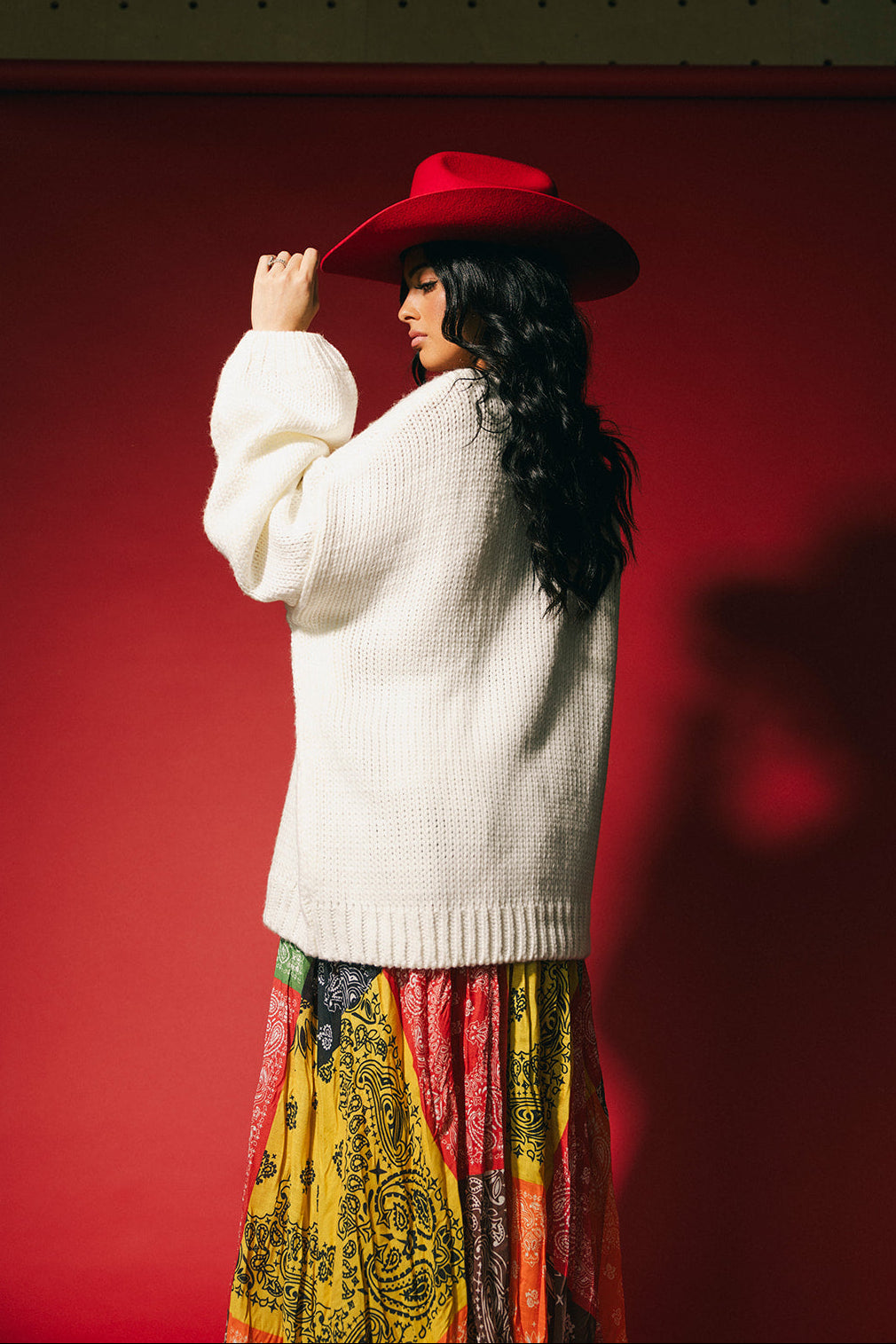 Person wearing a white sweater and colorful skirt with a red cowboy hat against a red background
