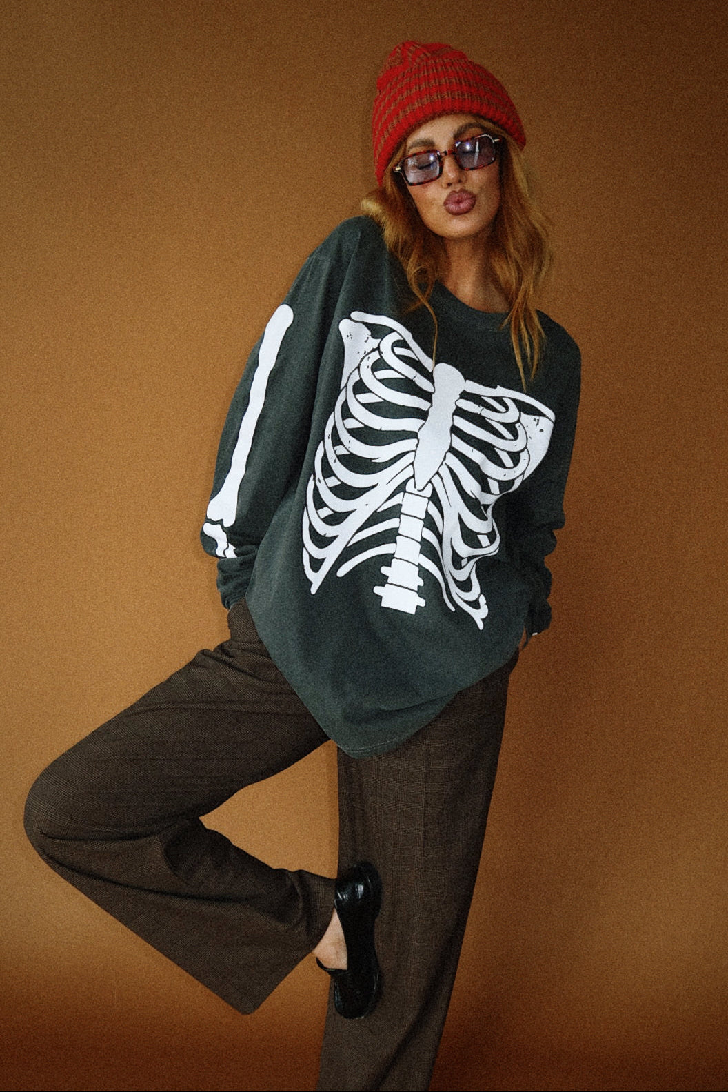 Person wearing a dark green sweater with a white skeleton design, brown pants, and a red beanie on a brown background.