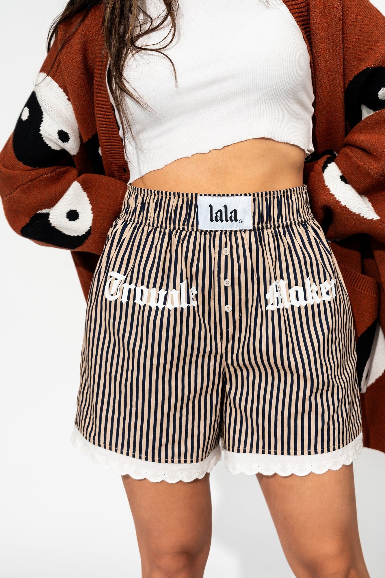 photo of girl wearing tan and navy stripe boxer shorts that say "trouble maker" across the front in white and has white lace trim on the hem with bows, elastic waistband, side pockets, 3 button faux fly, and white label that says "lala" on the waistband
