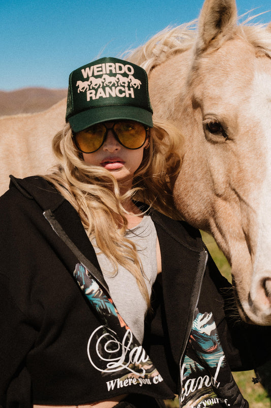 Person wearing a 'Weirdo Ranch' cap and sunglasses standing next to a horse.