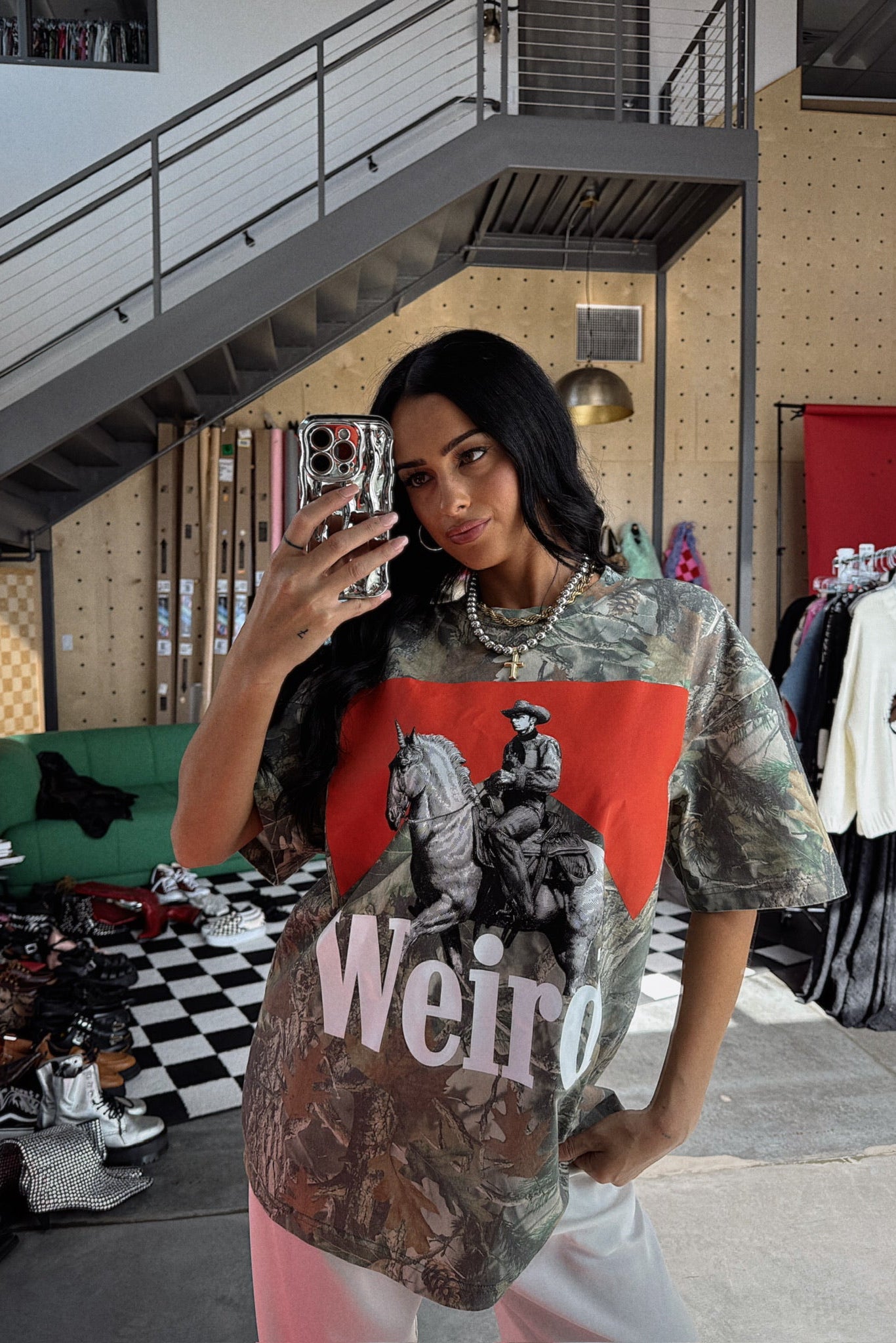 Woman taking a mirror selfie wearing a graphic t-shirt with a horse and 'Weird' text in a casual setting.