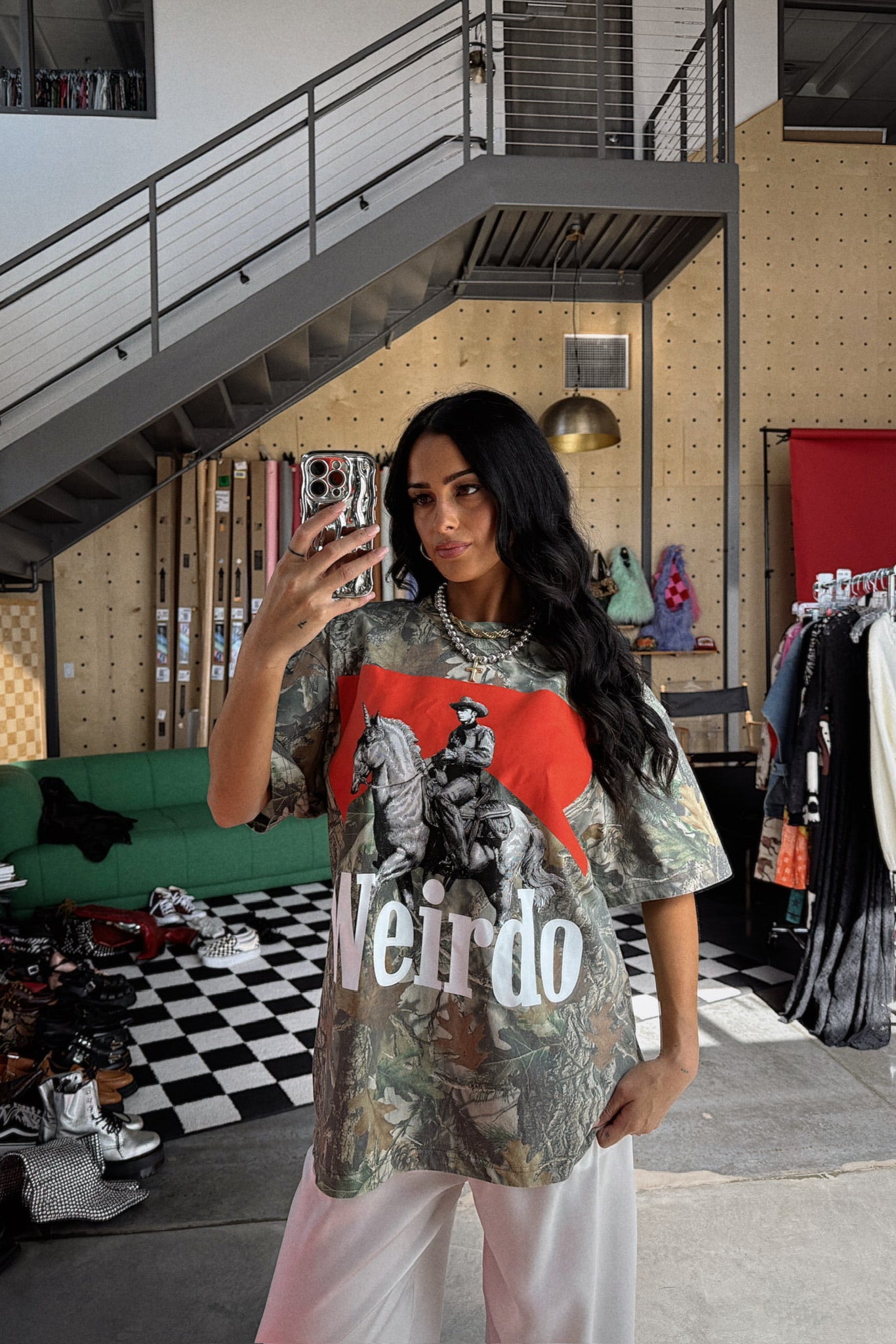 Person taking a mirror selfie wearing a graphic t-shirt in a room with a staircase and clothing rack.