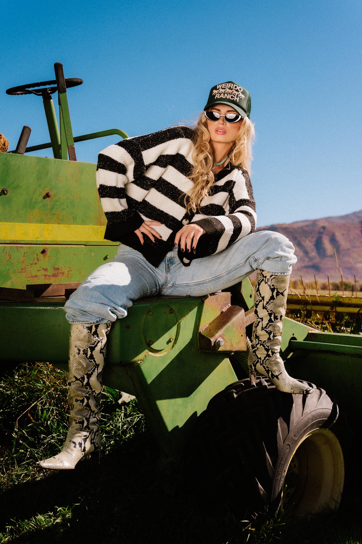 Person sitting on a green tractor wearing a black and white striped sweater, jeans, patterned boots, and green trucker hat with "WEIRDO RANCH" text and horses in puff print with a scenic background.