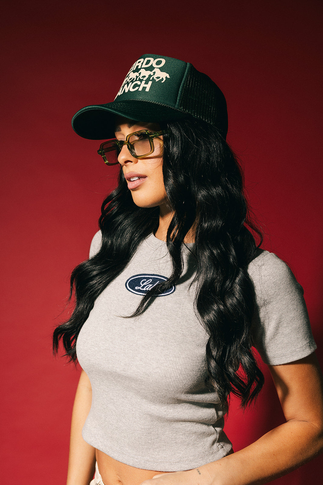 Woman wearing a green trucker hat with 'WEIRDO RANCH' text and horse graphics in puff print and gray top with a logo against a red background