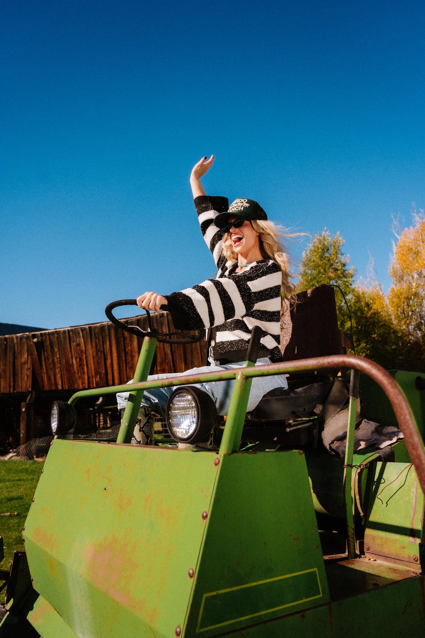 Person in a striped cardigan and green trucker hat with 'WEIRDO RANCH' text and horse graphics in puff print driving a green tractor with a clear blue sky in the background