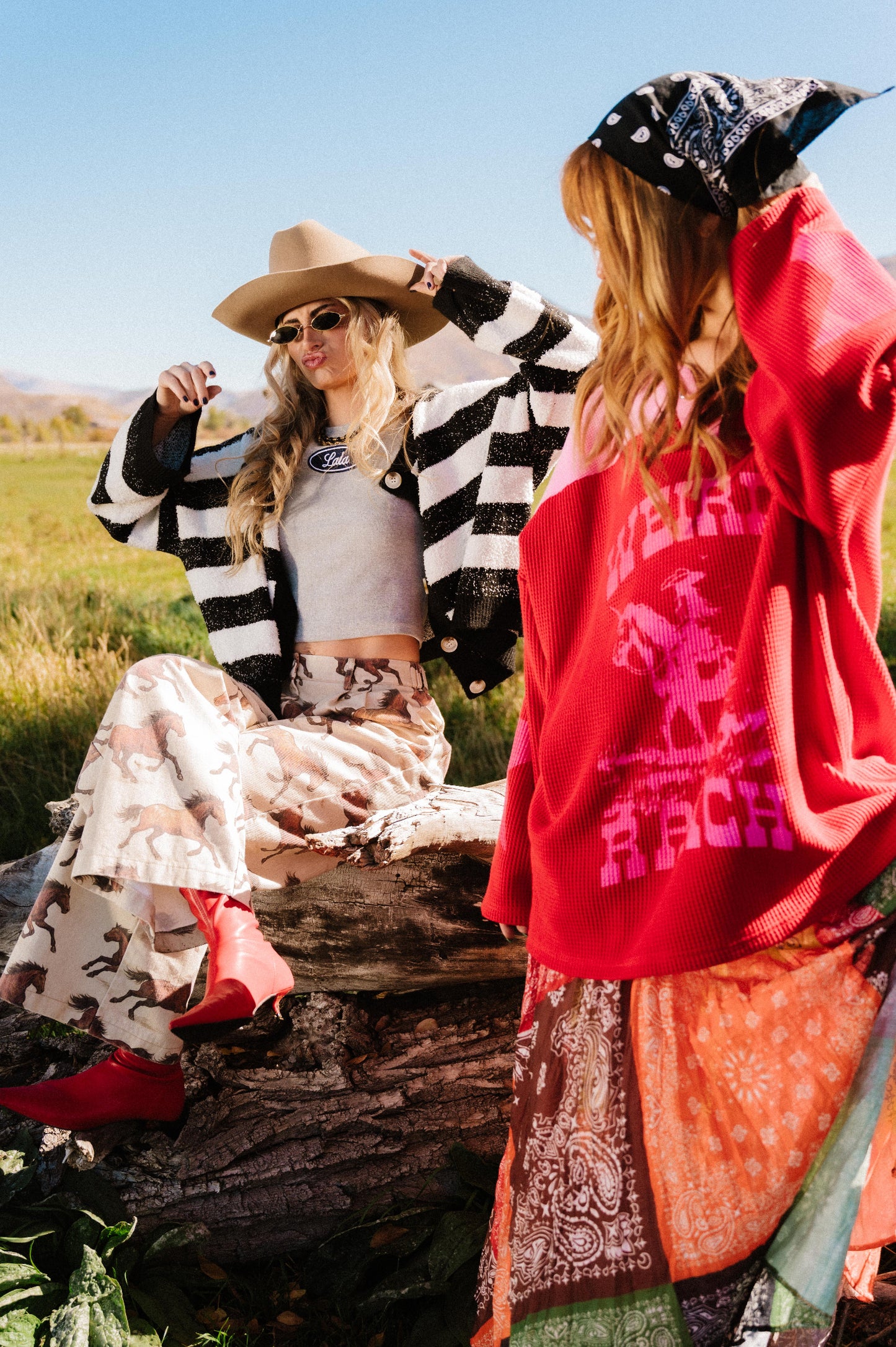 Two women sitting outdoors with one wearing a red and pink waffle knit hockey jersey inspired top with 'Weirdo Ranch' text and cowgirl riding a unicorn graphic.