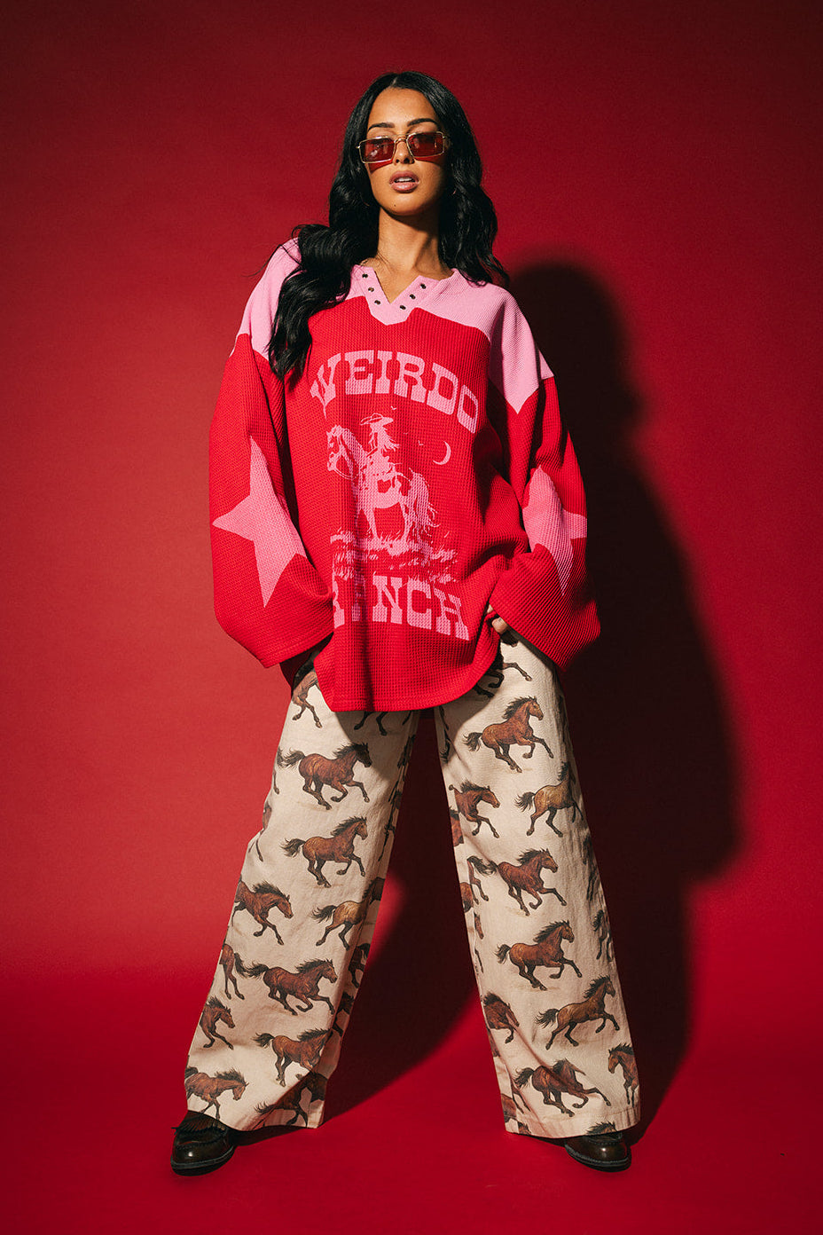 Person wearing a red and pink waffle knit hockey jersey inspired top with 'Weirdo Ranch' text and cowgirl riding a unicorn graphic on the front and star graphics on the sleeves on a red background