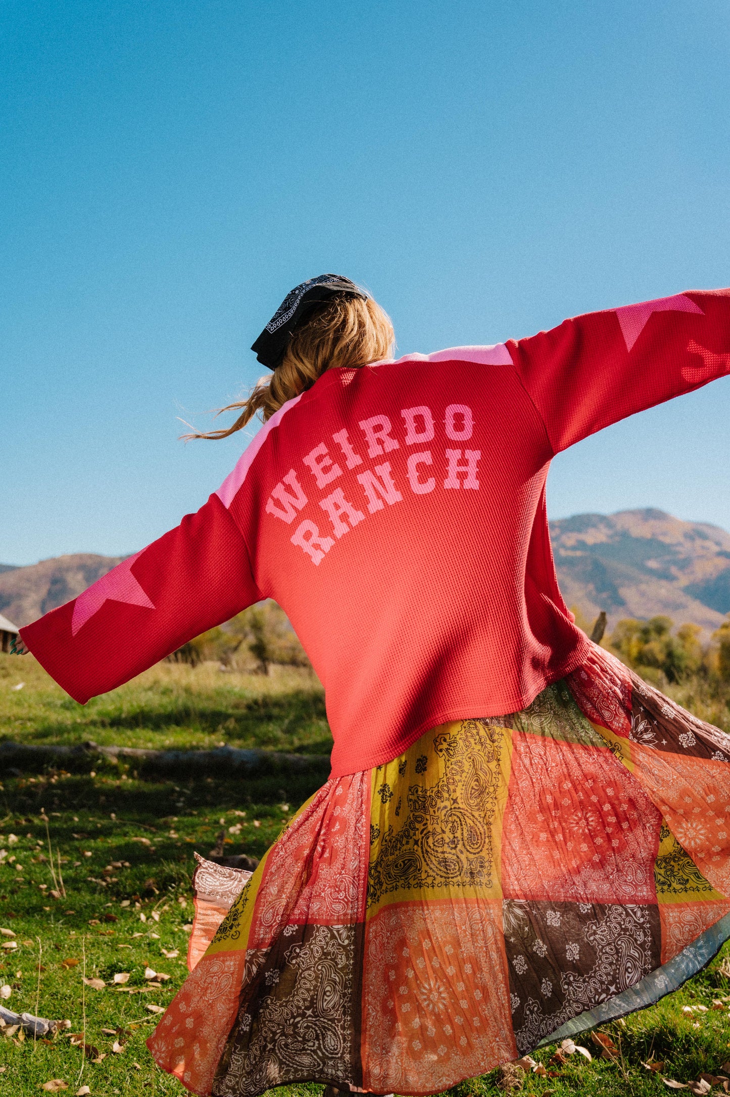 Person wearing a red and pink hockey jersey inspired top with 'WEIRDO RANCH' text with star graphics on the sleeves, standing in a field with mountains in the background.