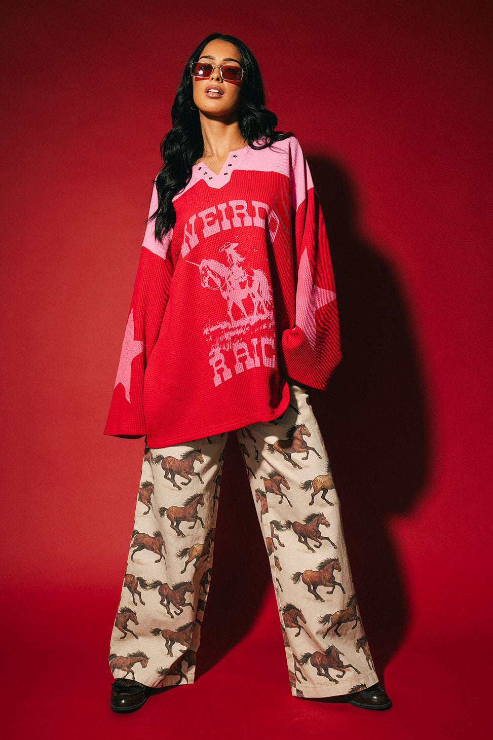 Person wearing a red and pink waffle knit hockey jersey inspired top with 'Weirdo Ranch' text and cowgirl riding a unicorn graphic on the front and star graphics on the sleeves on a red background.