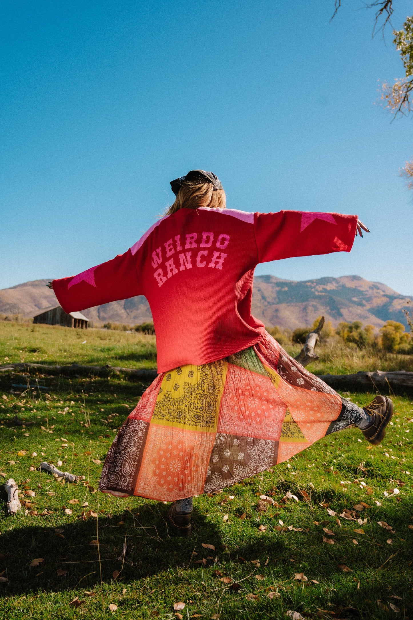 Person wearing a red and pink waffle knit hockey jersey inspired top with 'Weirdo Ranch' text on the back and star graphics on the sleeves standing in a grassy field with mountains in the background.