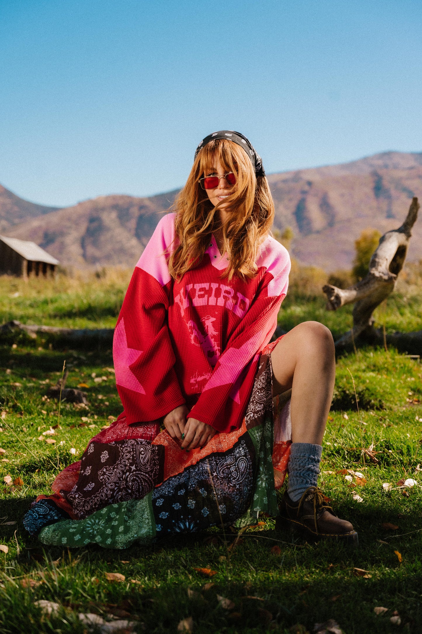 Person sitting on grass wearing a red and pink waffle knit hockey jersey inspired top with 'Weirdo Ranch' text and cowgirl riding a unicorn graphic on the front and star graphics on the sleeves.