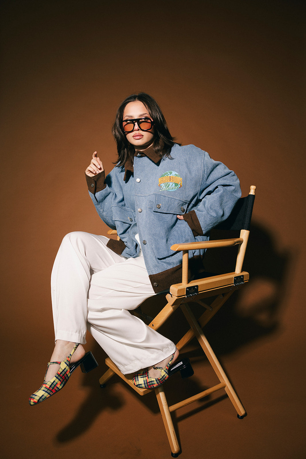 Person sitting on a director's chair wearing a blue denim jacket and sunglasses against a brown background