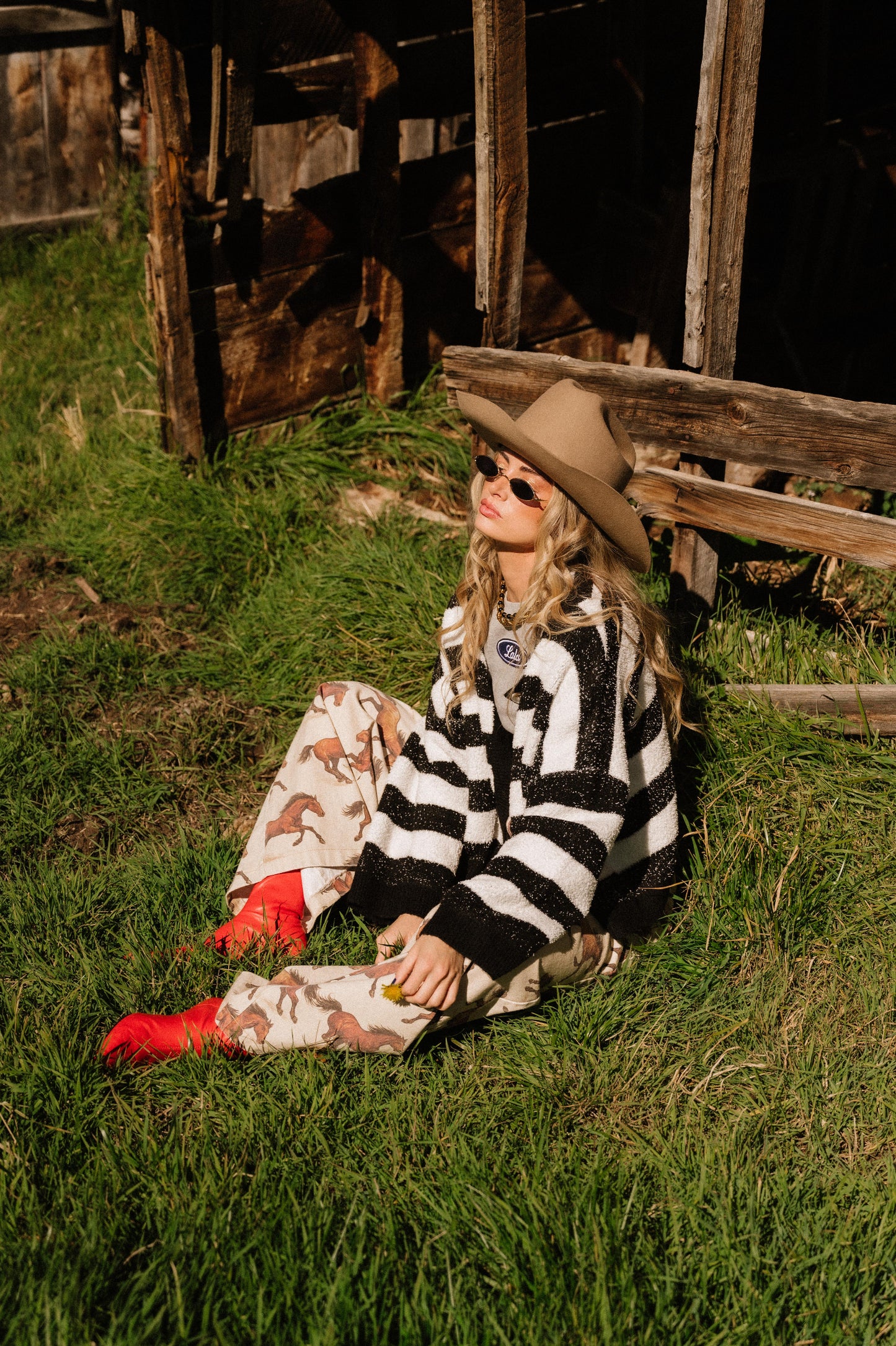 Person sitting on grass wearing a black and white striped boxy sweater and sunglasses, with a rustic wooden structure in the background.