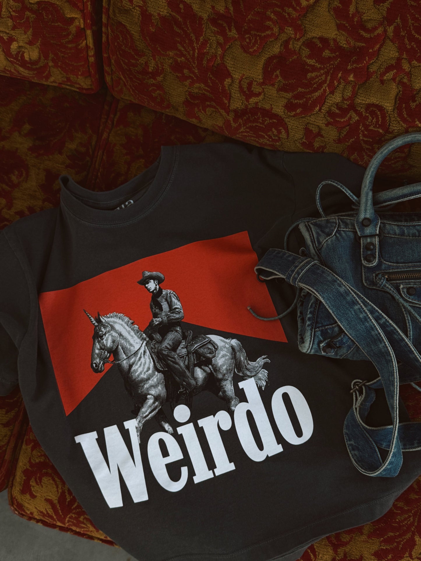 Black t-shirt with a graphic of a person on a horse and the word 'Weirdo' on a red background, placed on a patterned surface.