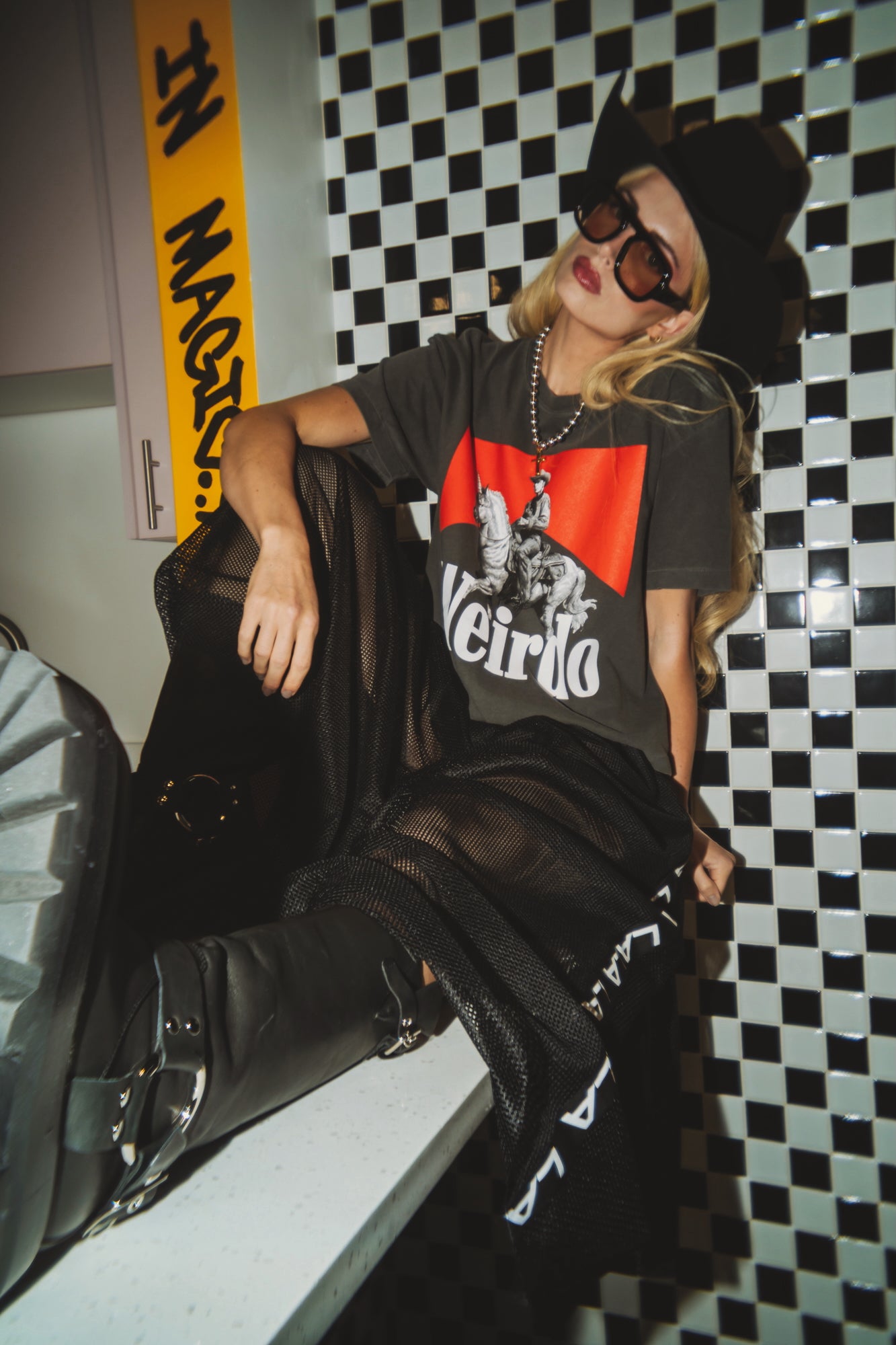 Person wearing a black t-shirt with a graphic design, sitting against a checkered wall.