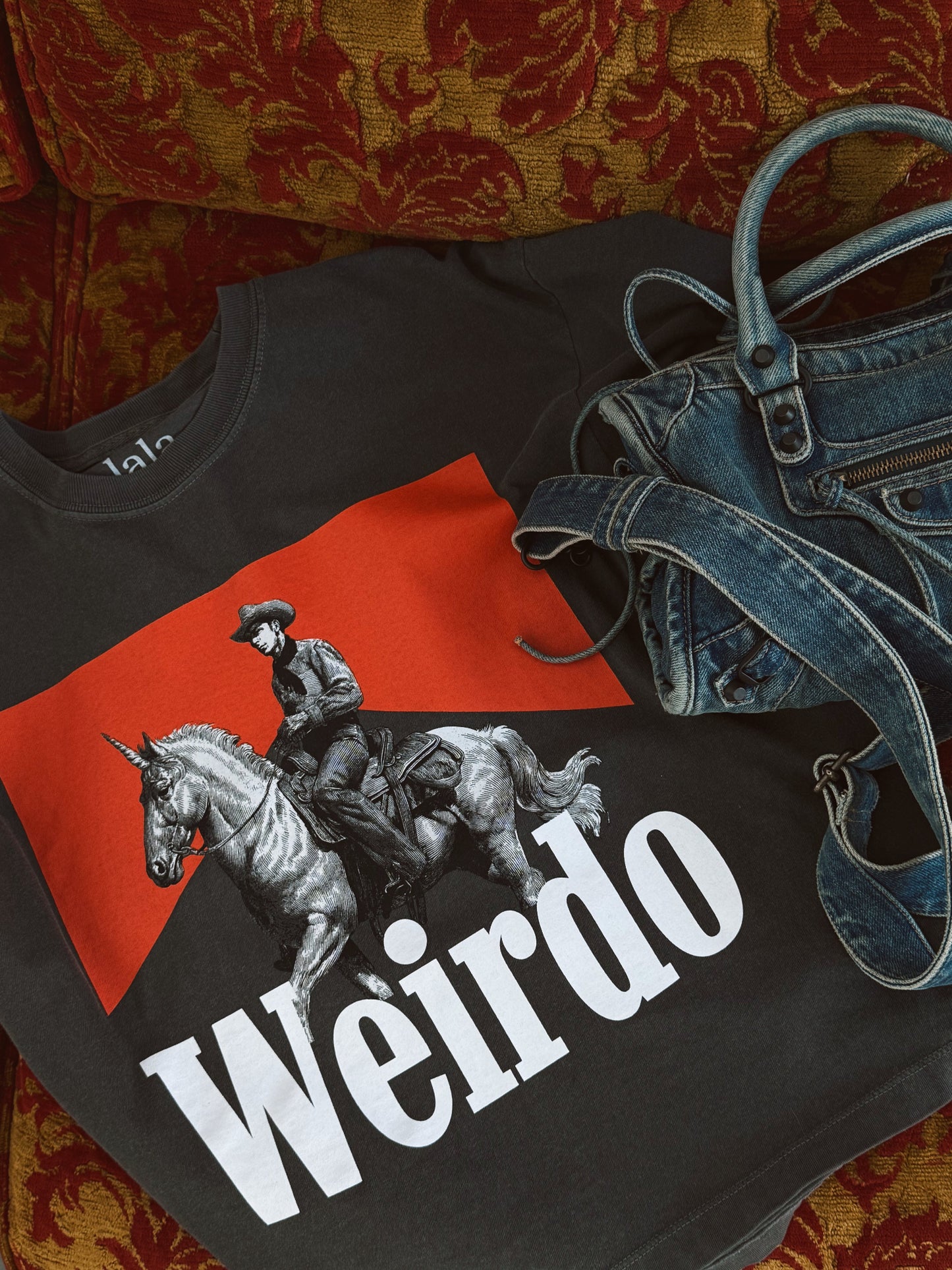 Black t-shirt with red and white graphic design featuring a person on a horse and the word 'Weirdo', placed on a patterned surface with jeans.