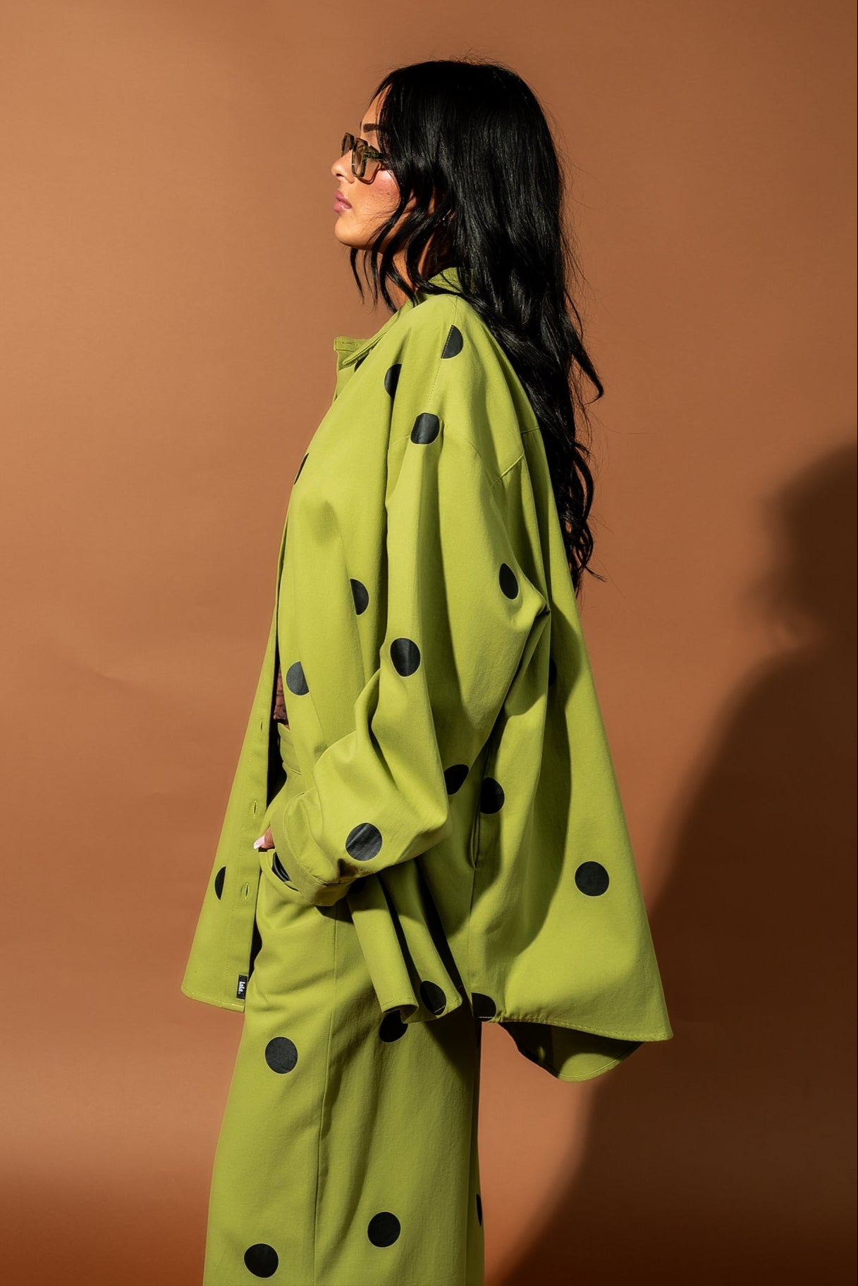 Girl/model standing against a brown backdrop wearing a green shirt jacket with black polka dots and matching pants