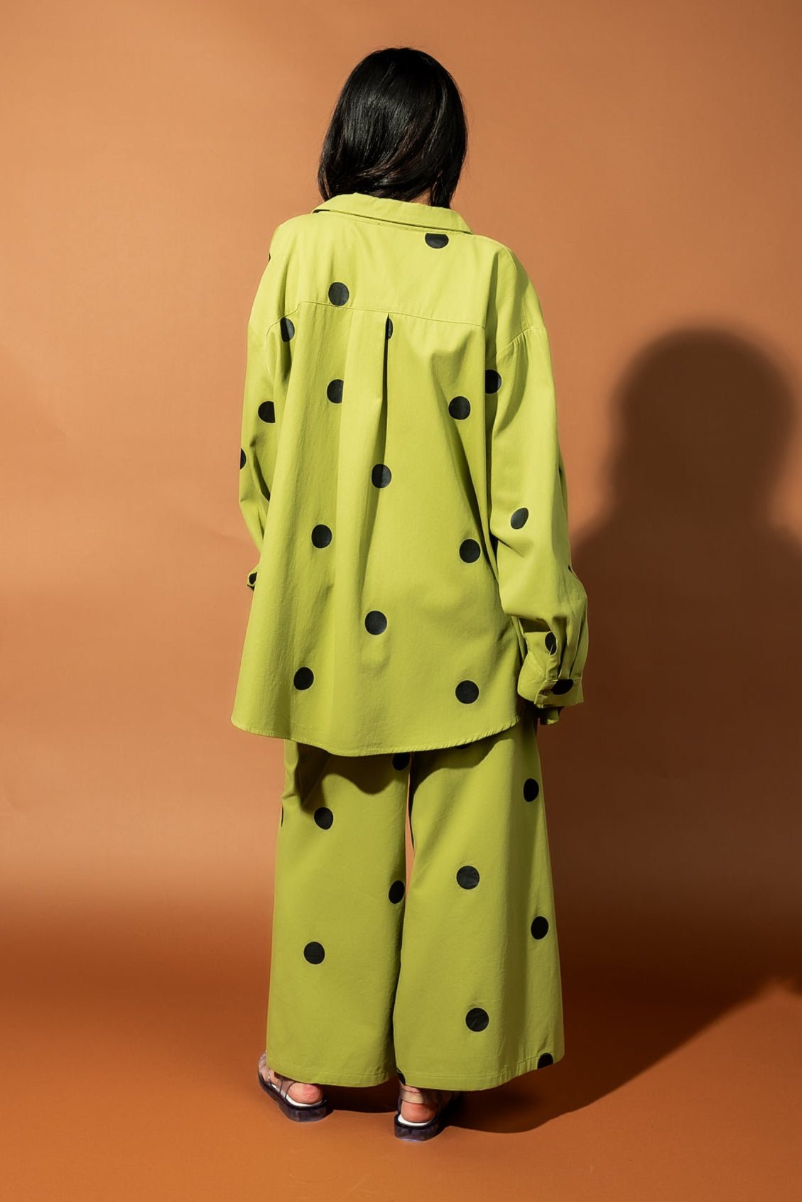 Girl/model standing against a backdrop facing backwards wearing a green shirt jacket with black polka dots and matching pants