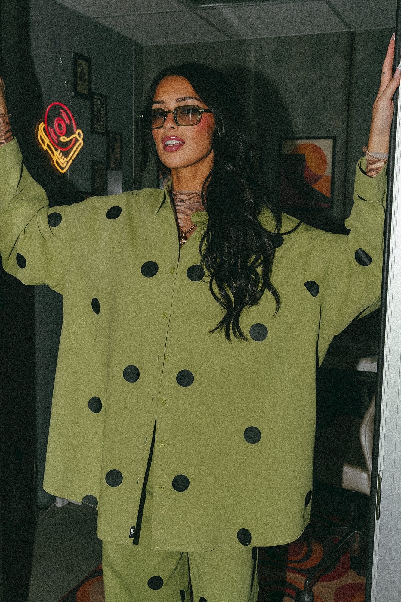 Girl/model standing against a door frame wearing a green shirt jacket with black polka dots and matching pants