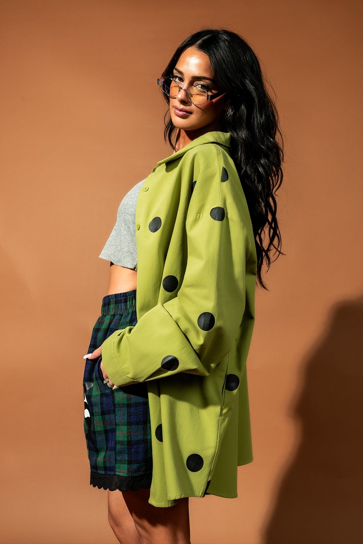 Girl/model standing against a brown backdrop wearing a green shirt jacket with black polka dots, blue and green flannel boxers and a grey tank top