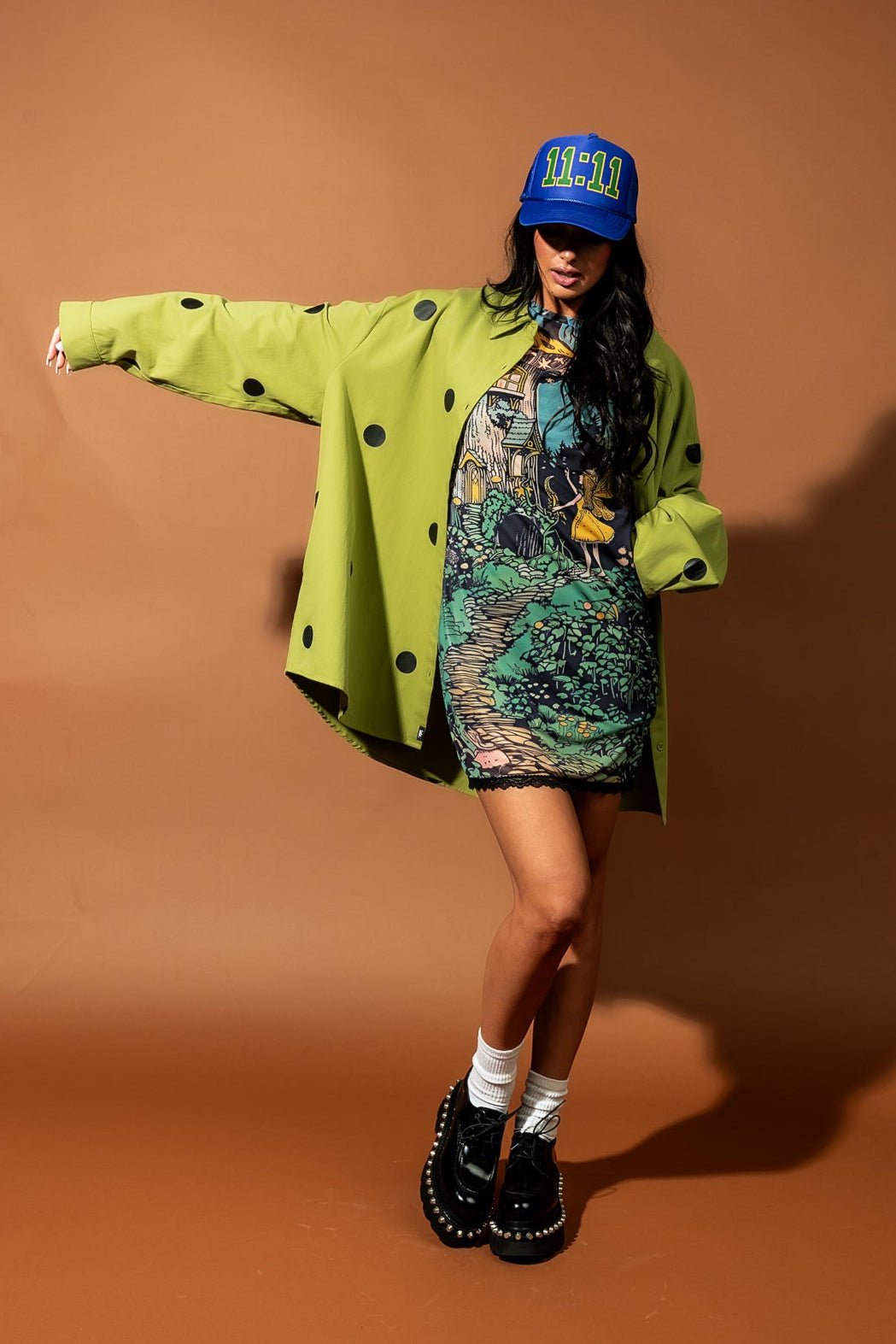 Girl/model standing against a brown backdrop wearing a green shirt jacket with black polka dots, a mesh dress with a fairy pattern on it and a blue trucker hat with 11:11 graphics