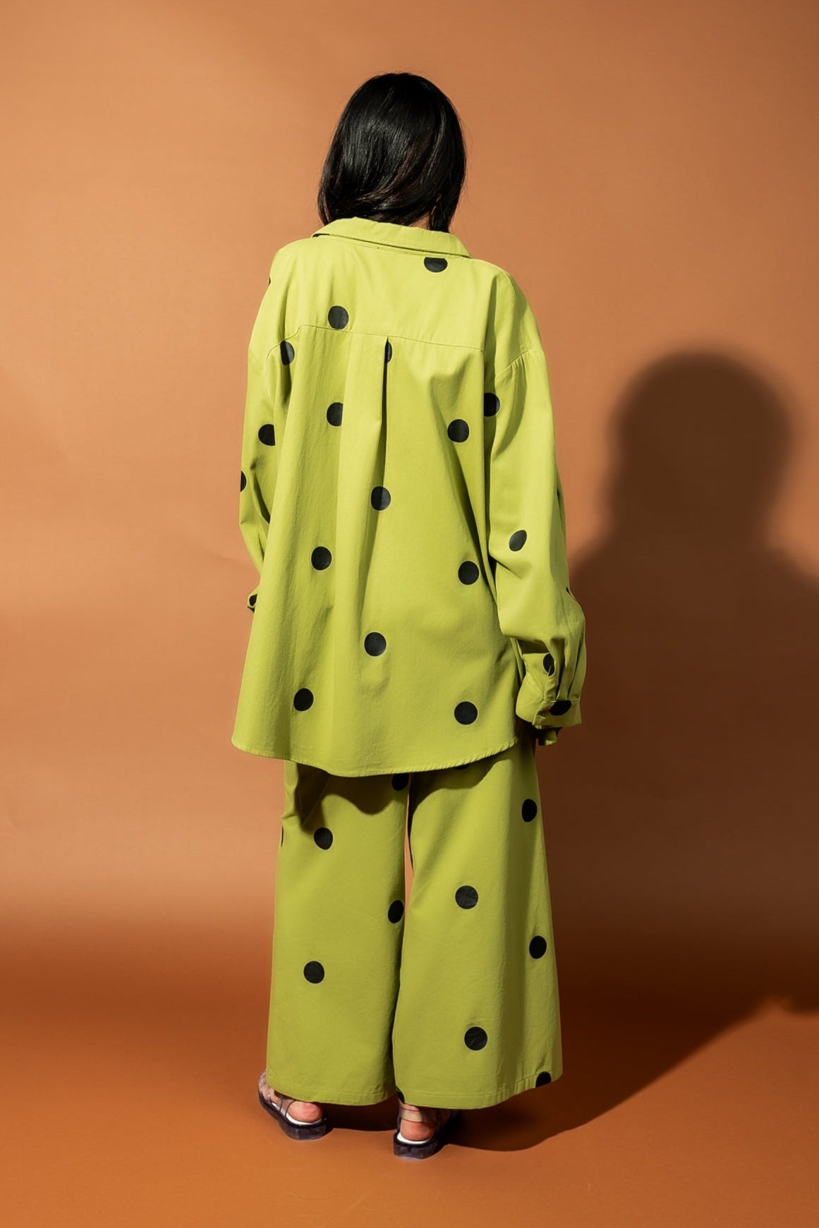 Girl/model standing against a brown backdrop wearing a green shirt jacket with black polka dots and matching pants, facing away