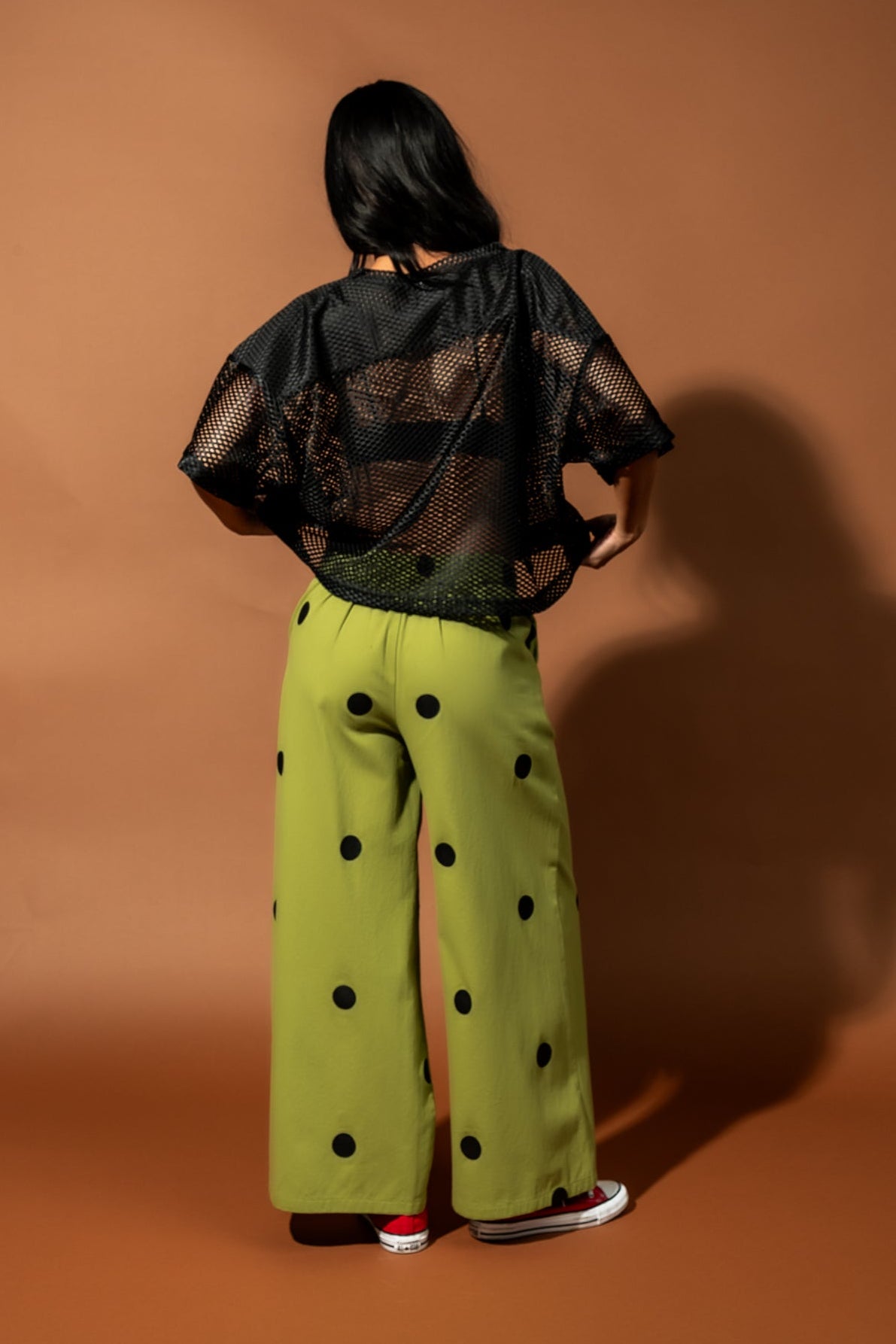 Girl/model standing against a brown backdrop wearing green pants with black polka dots and a black football jersey, facing away