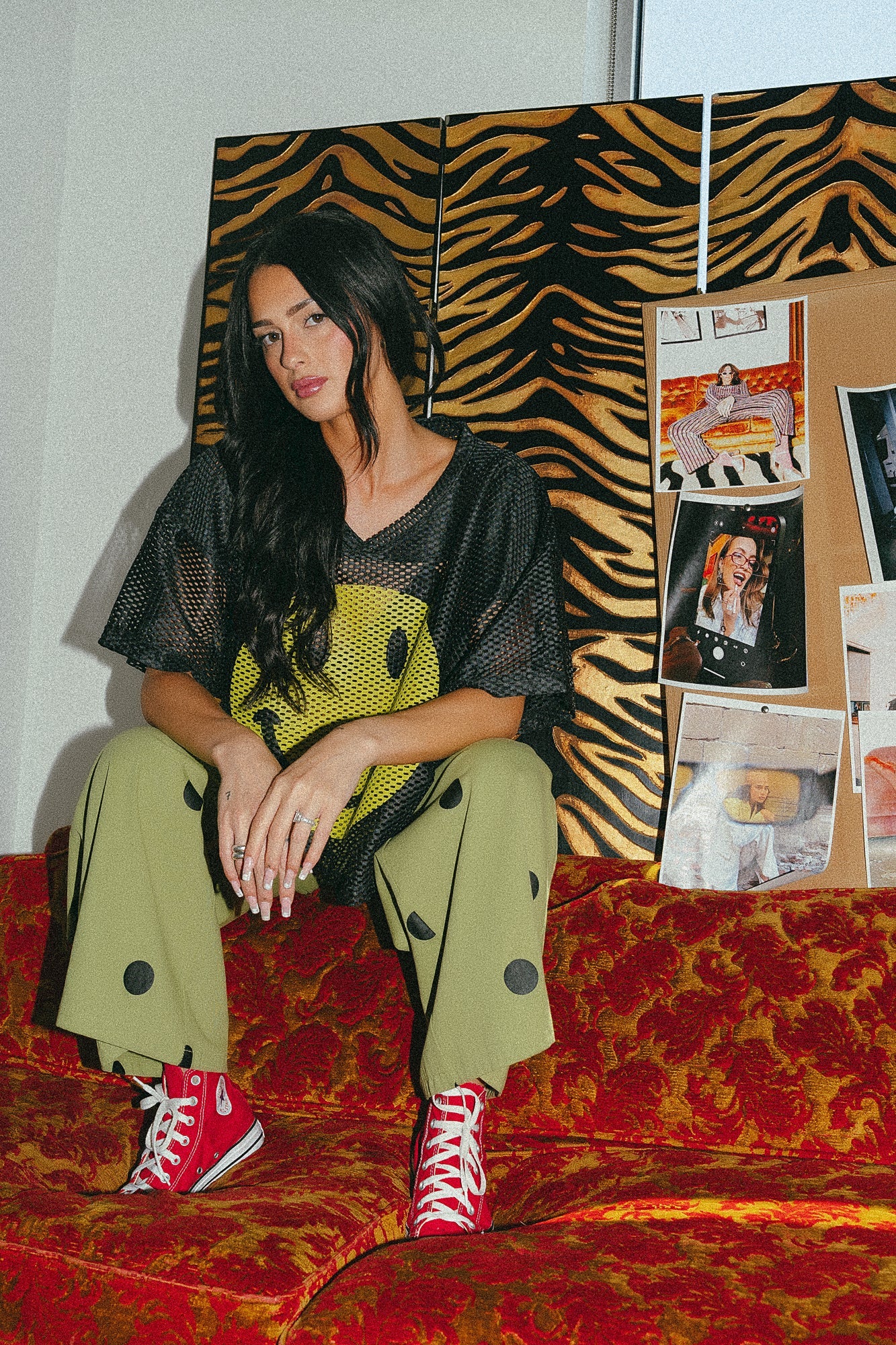 Photo of a girl/model sitting on a couch wearing green pants with black polka dots and a black football jersey with a big yellow smile face