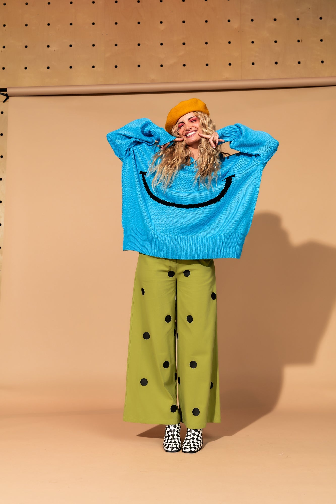 Girl/model standing against a brown backdrop wearing green pants with black polka dots and a blue sweater with a big black smile face on it