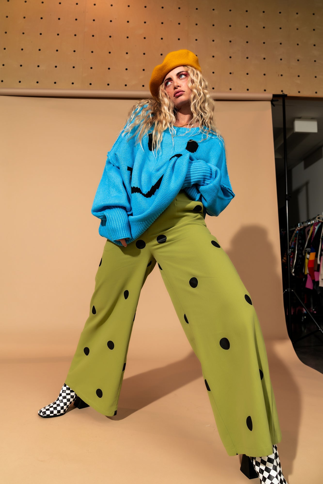 Girl/model standing against a brown backdrop wearing green pants with black polka dots, a blue sweater with a black smile face on it and a yellow beret