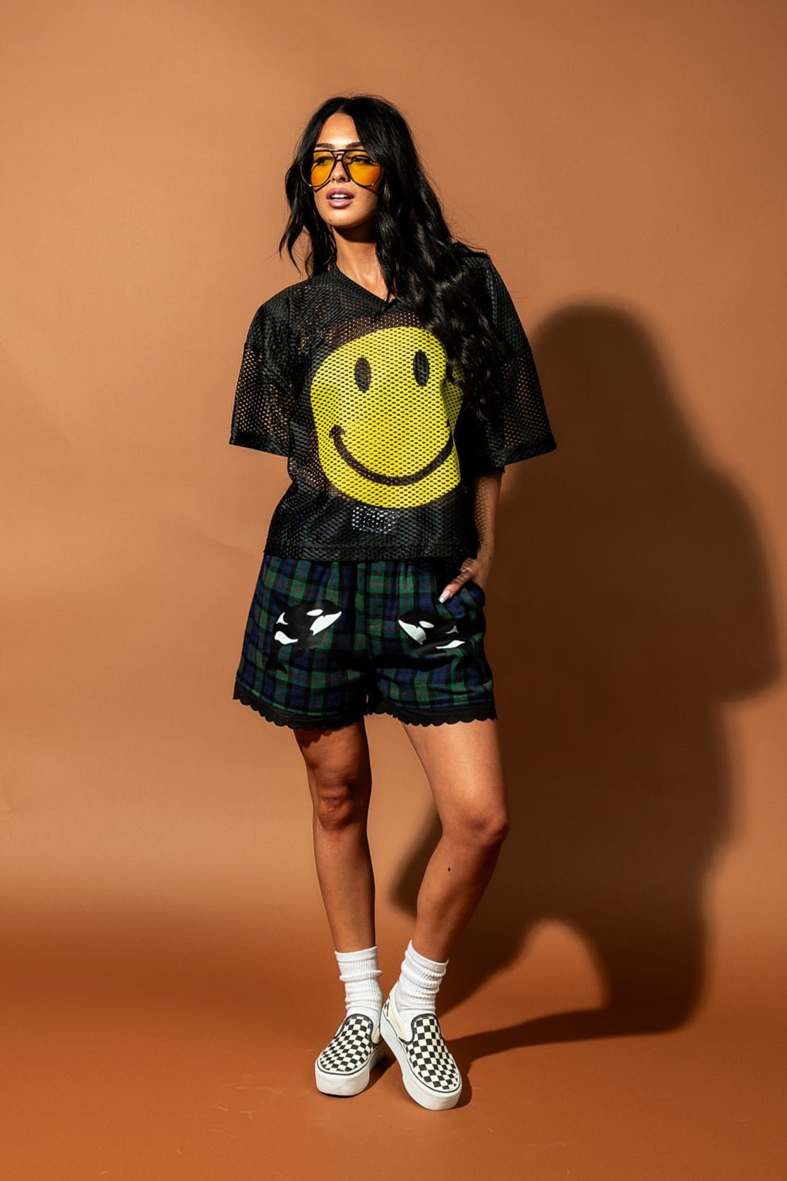 Girl/model wearing a black football jersey with a big yellow smile face printed on it with blue and green plaid shorts