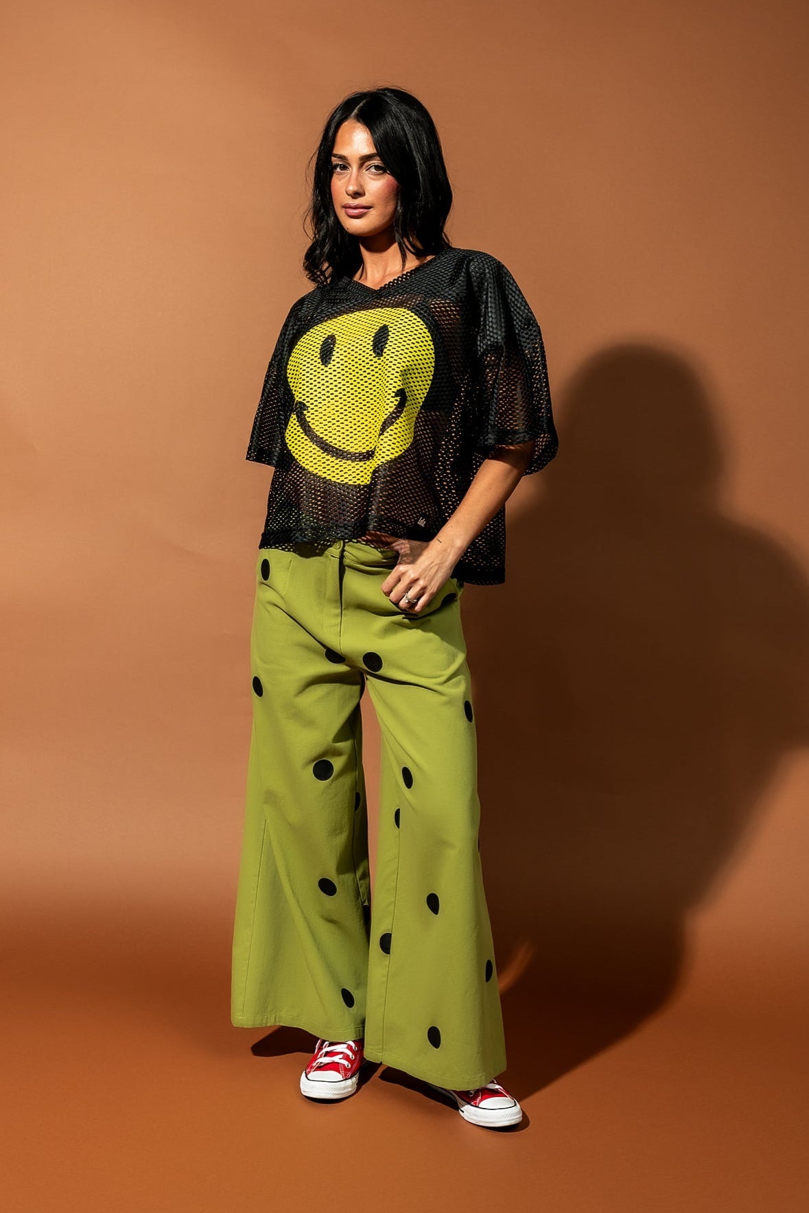 Girl/model wearing a black football jersey with a big yellow smile face printed on it with a pair of green pants with black polka dots