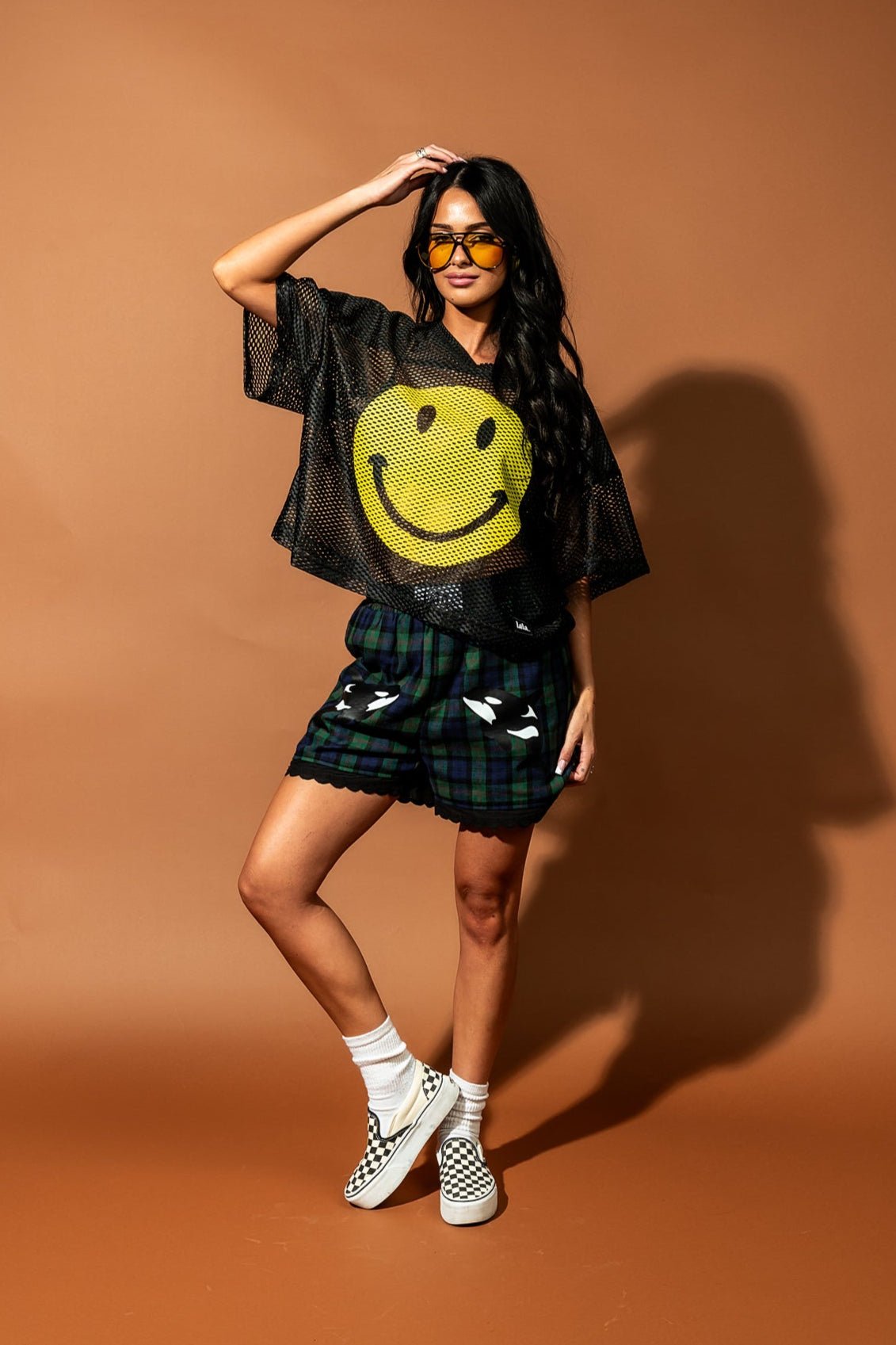 Girl/model wearing a black football jersey with a big yellow smile face printed on it with blue and green plaid shorts